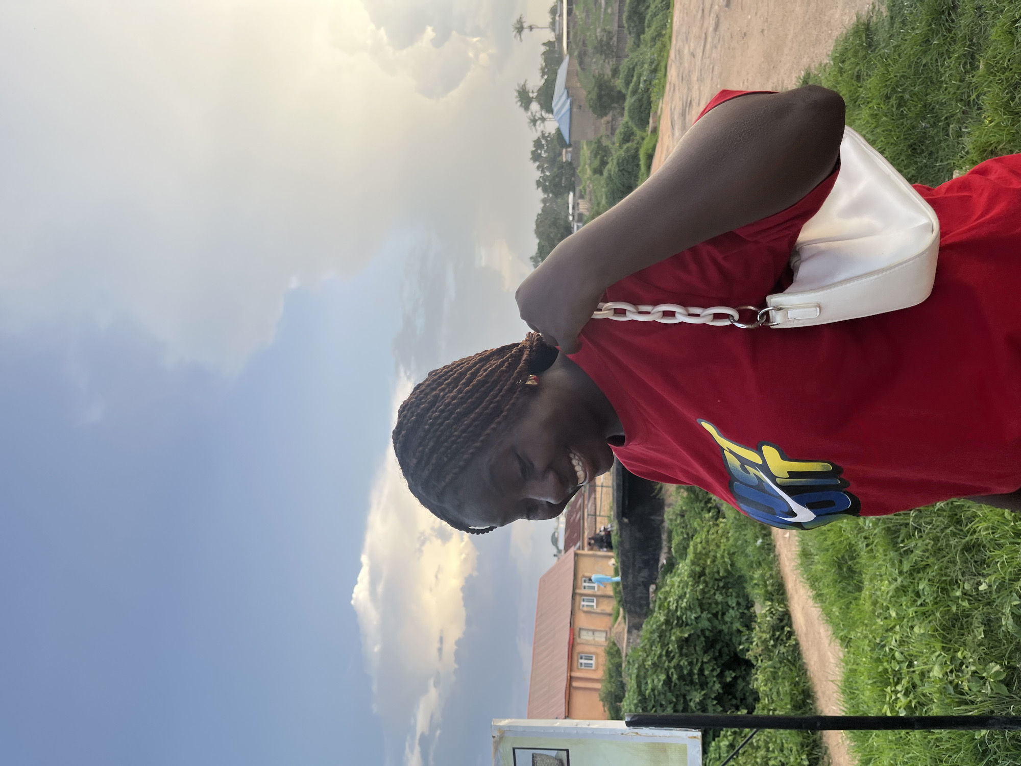 A young Black woman with braided hair smiles broadly while looking down. She is wearing a red t-shirt with a colorful "JUST DO IT" Nike logo on it and a white purse with a thick white chain strap is slung over her shoulder. The background is an outdoor scene with a dirt path, lush green grass, and trees. In the distance, there are buildings with orange and blue roofs, and a cloudy sky with a hint of sunlight. The overall setting appears to be a rural or suburban area.