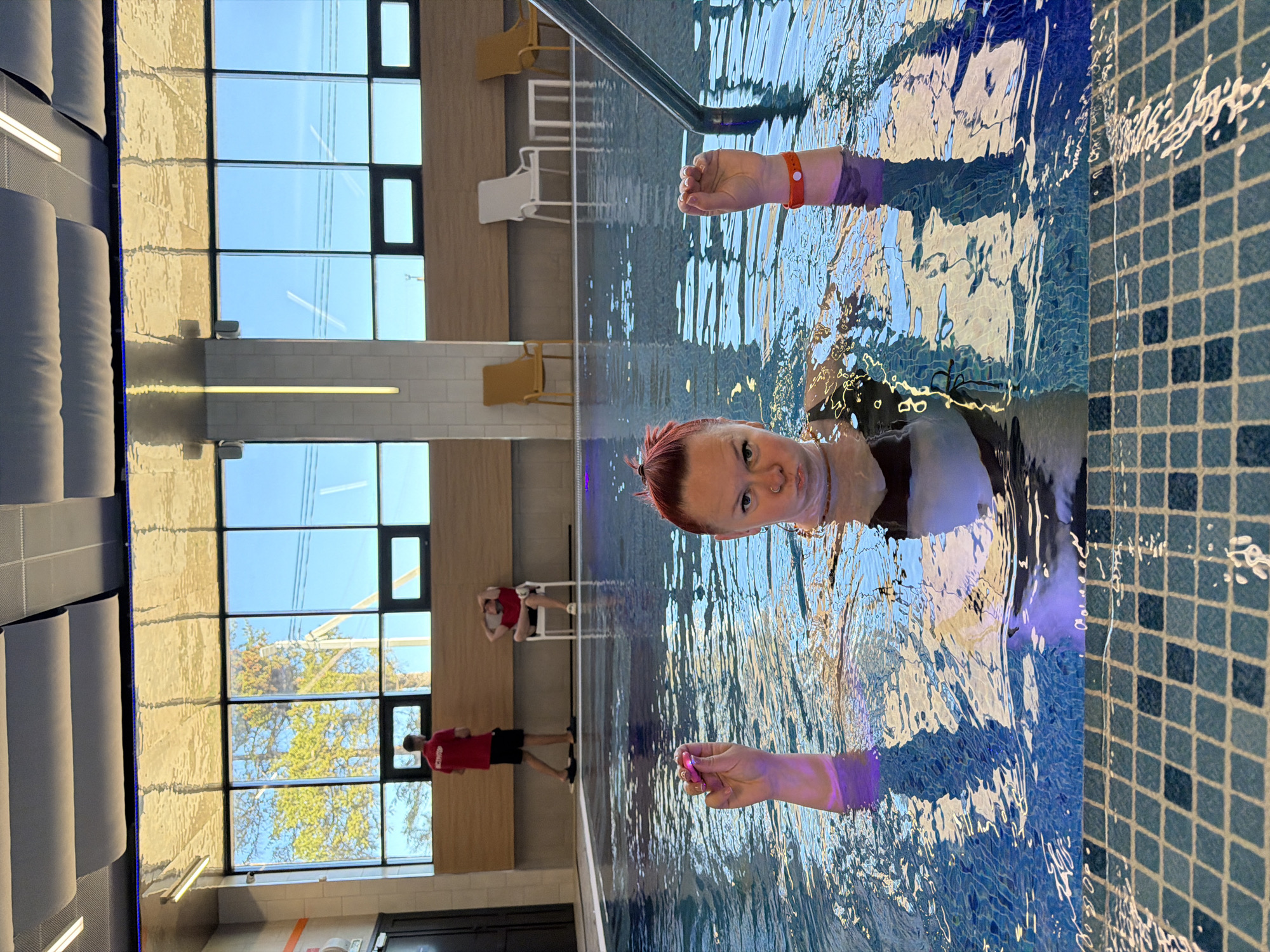 The image shows a person in a swimming pool. The person is partially submerged, with their head and shoulders above the water. The water is clear and blue, with visible ripples and reflections. The pool has a tiled border, and there is a handrail along the side. In the background, there is a wall with large windows that are reflecting the sky and some trees. On the wall, there are chairs, and there appear to be two people in red shirts, one standing and one sitting. The location is Votkinsk, Russia.