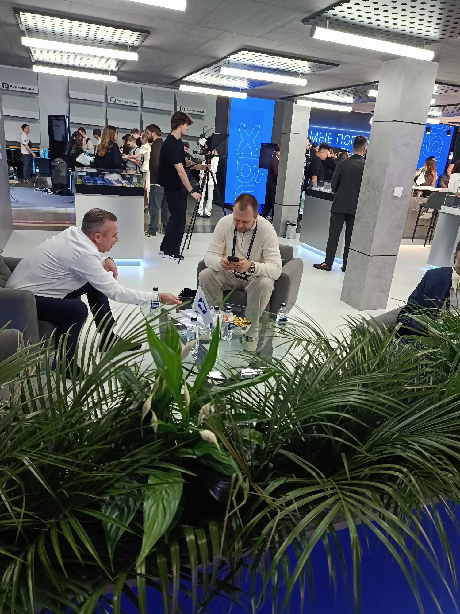 The scene is set in what appears to be an exhibition hall or a large open office space. A number of people are present, engaged in various activities. In the foreground, two men are seated in chairs around a glass coffee table covered with water bottles and snacks. One man, dressed in a white shirt and dark trousers, is gesturing towards the table, while the other, wearing a beige sweater, is looking at his phone. Large potted plants are positioned in front of them, partially obscuring the view.
In the background, other people are milling around, some standing at displays or counters, and others walking through the space. There's a cameraman with a tripod set up, possibly recording the event. The environment includes overhead lighting panels and large pillars that seem to be part of the building's structure. The scene is located in the city of Unknown, Unknown.