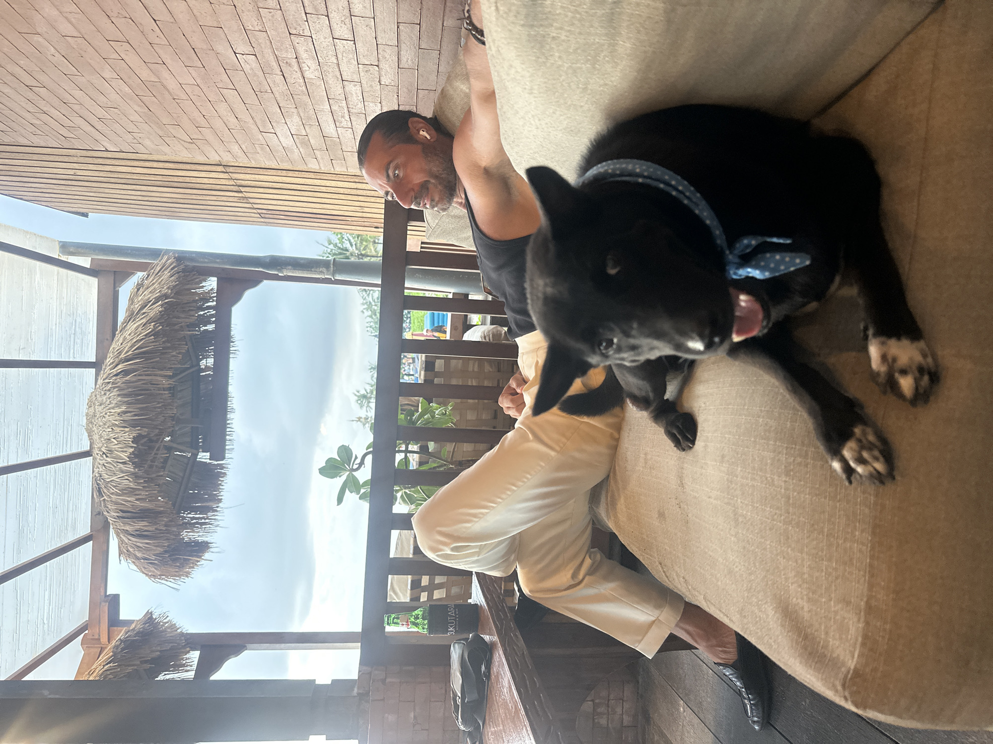 A man with dark hair and a beard is sitting on a cushioned sofa outdoors in North Kuta, Indonesia. The man is wearing a black tank top and light-colored pants. A black dog with white markings on its paws and a blue polka dot collar is lying on the sofa next to him. The dog's tongue is sticking out. The setting appears to be a patio or balcony with a thatched roof structure visible above. A wooden railing is in the background, with plants visible beyond. In front of the man is a table that is holding a drink and other personal items. The man is looking at the dog and smiling.
