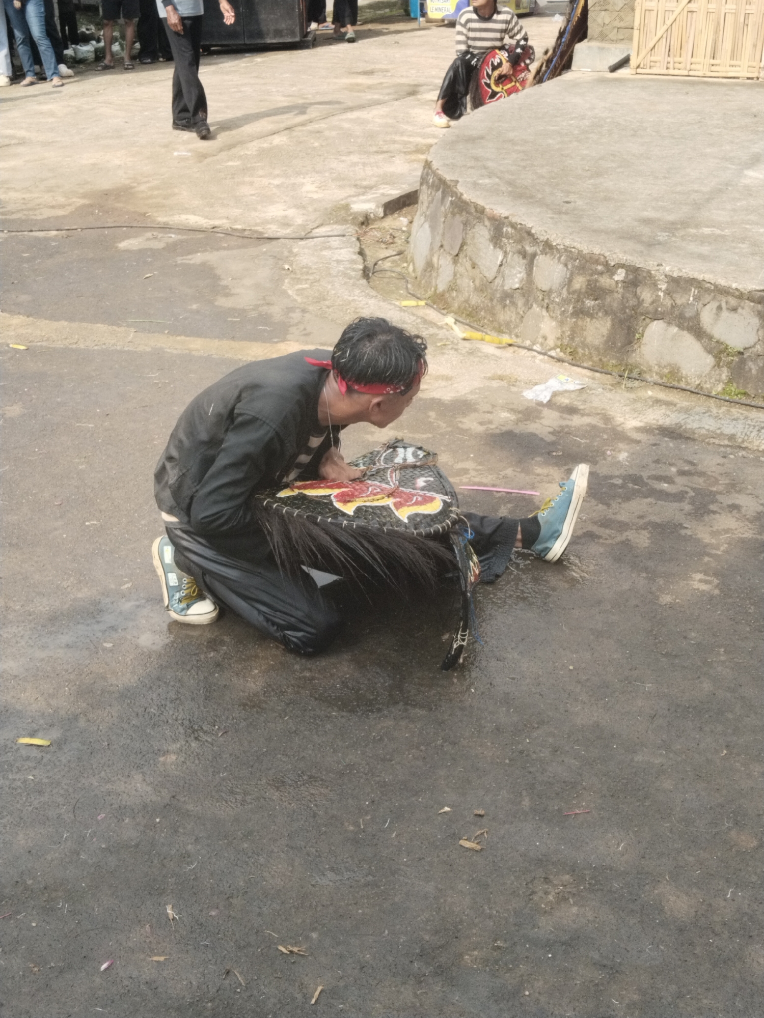 The scene depicts a man kneeling on a paved surface, possibly a street or courtyard, in Pendem, Indonesia. The man, wearing a black jacket, black pants, a red headband, and blue sneakers, is hunched over a large, black and red woven object with horsehair-like fringe. The object appears to be a decorative mask. A few individuals are visible in the background, along with a speaker and a small structure made of bamboo. The ground is slightly wet.