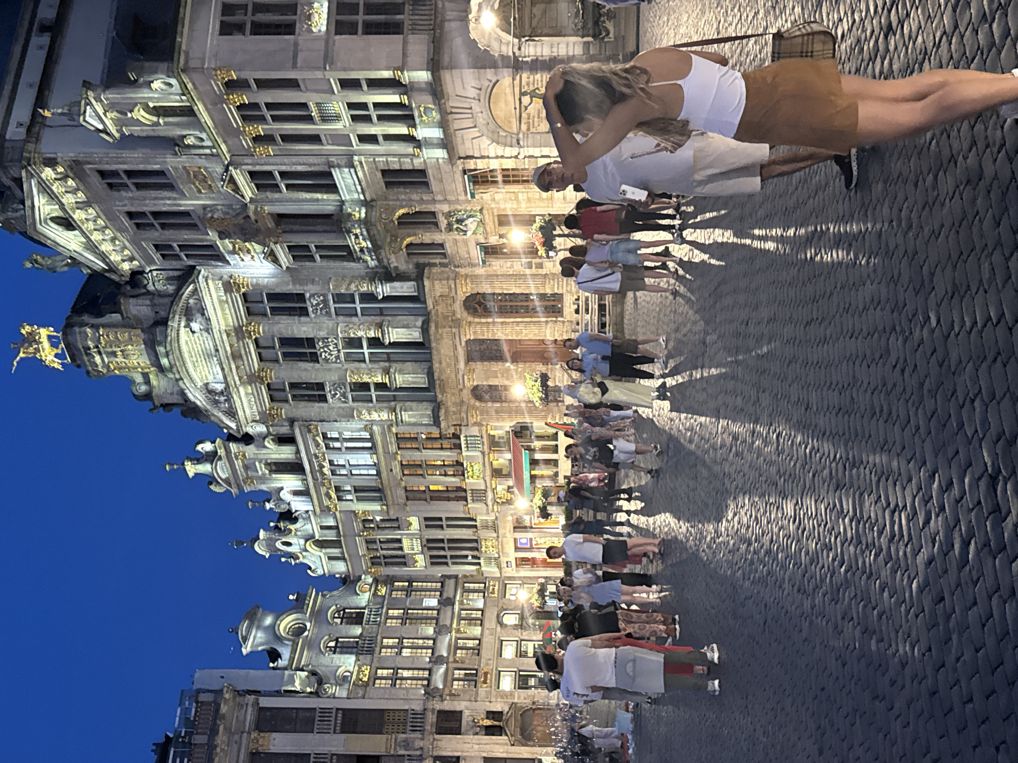 The image captures a nighttime scene in the Grand Place in Brussels, Belgium. Ornate, historic buildings with gold accents and numerous windows are illuminated against a dark blue sky. A crowd of people are gathered in the cobblestone square, with some walking and others standing and conversing. The lighting from the buildings casts long shadows across the ground. In the foreground, a woman with blonde hair stands with her back to the camera, her hand touching her hair. She is wearing a white tank top and a brown skirt, with a plaid shoulder bag. Other individuals in the foreground and middle ground are seen in casual attire, suggesting a tourist or evening outing. The overall atmosphere is one of a bustling public space at dusk or early evening.