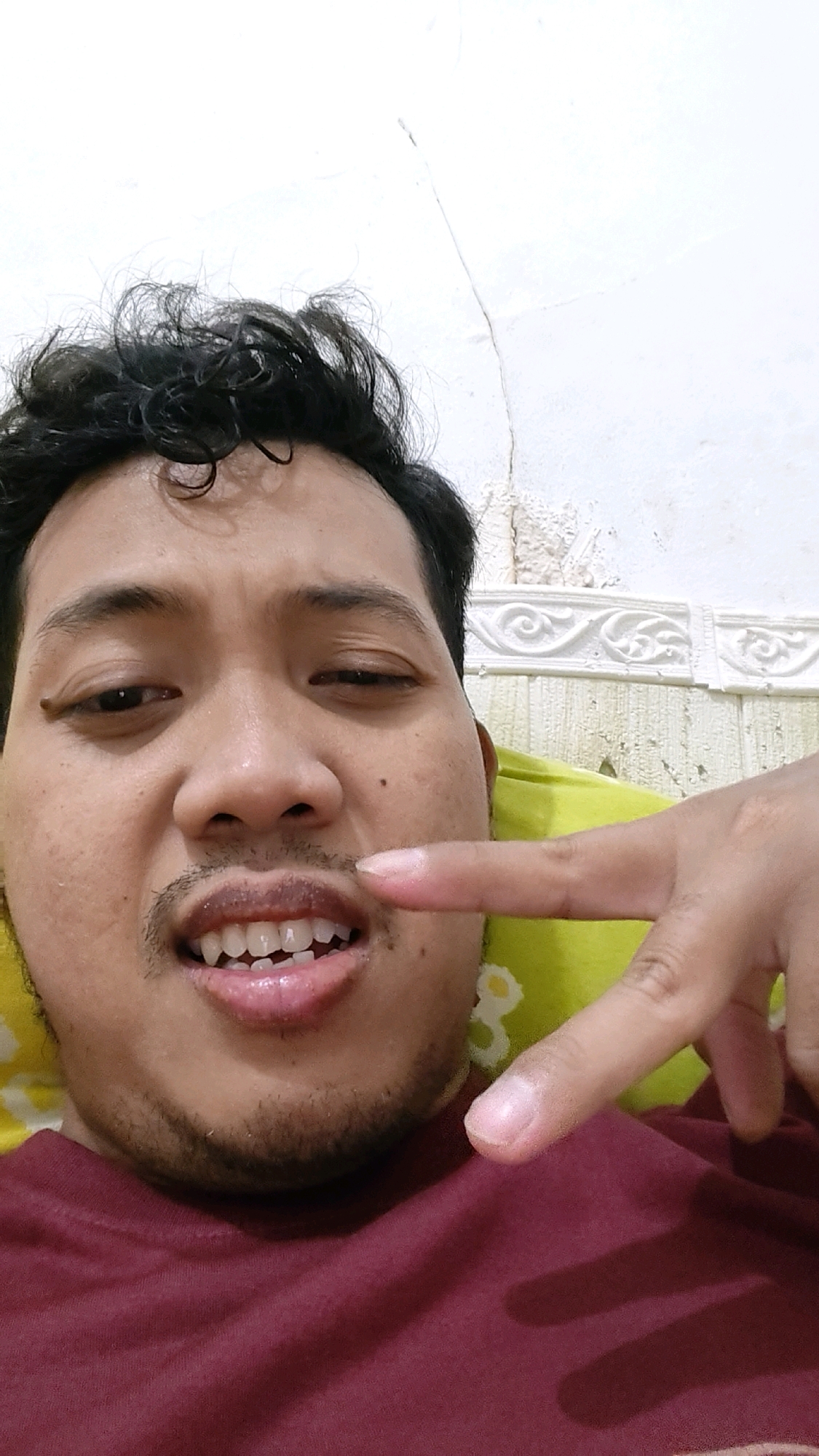 In Mekarmukti, Indonesia, a close-up selfie captures a man with dark, curly hair against a light-colored wall with decorative molding. He is wearing a maroon t-shirt and is lying down, possibly on a yellow pillow, as indicated by a portion of it visible. The man is winking and pointing his index finger towards his mouth, which is slightly open, revealing his teeth. He has a faint mustache and stubble on his chin and jawline. The lighting suggests an indoor setting.