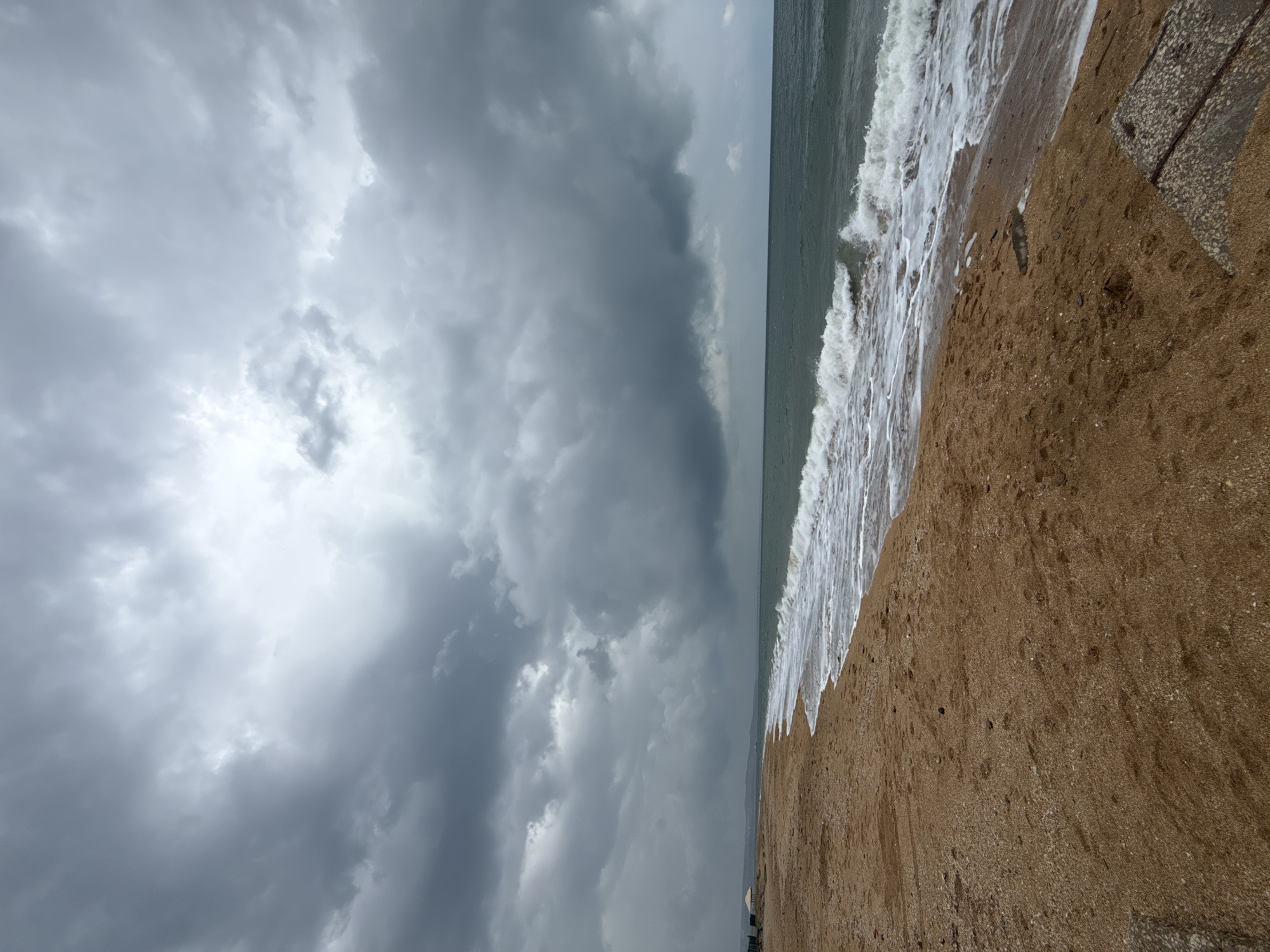A sandy beach on a cloudy day. The sky is filled with dark, heavy clouds, suggesting an impending storm. The sea is a deep green, with white-capped waves rolling towards the shore. The sand is a light brown color, with some scattered shells and pebbles. In the distance, there is a faint line of trees and buildings. The image captures the raw beauty of nature and the power of the elements. The location is the beach of Kerch, Ukraine.