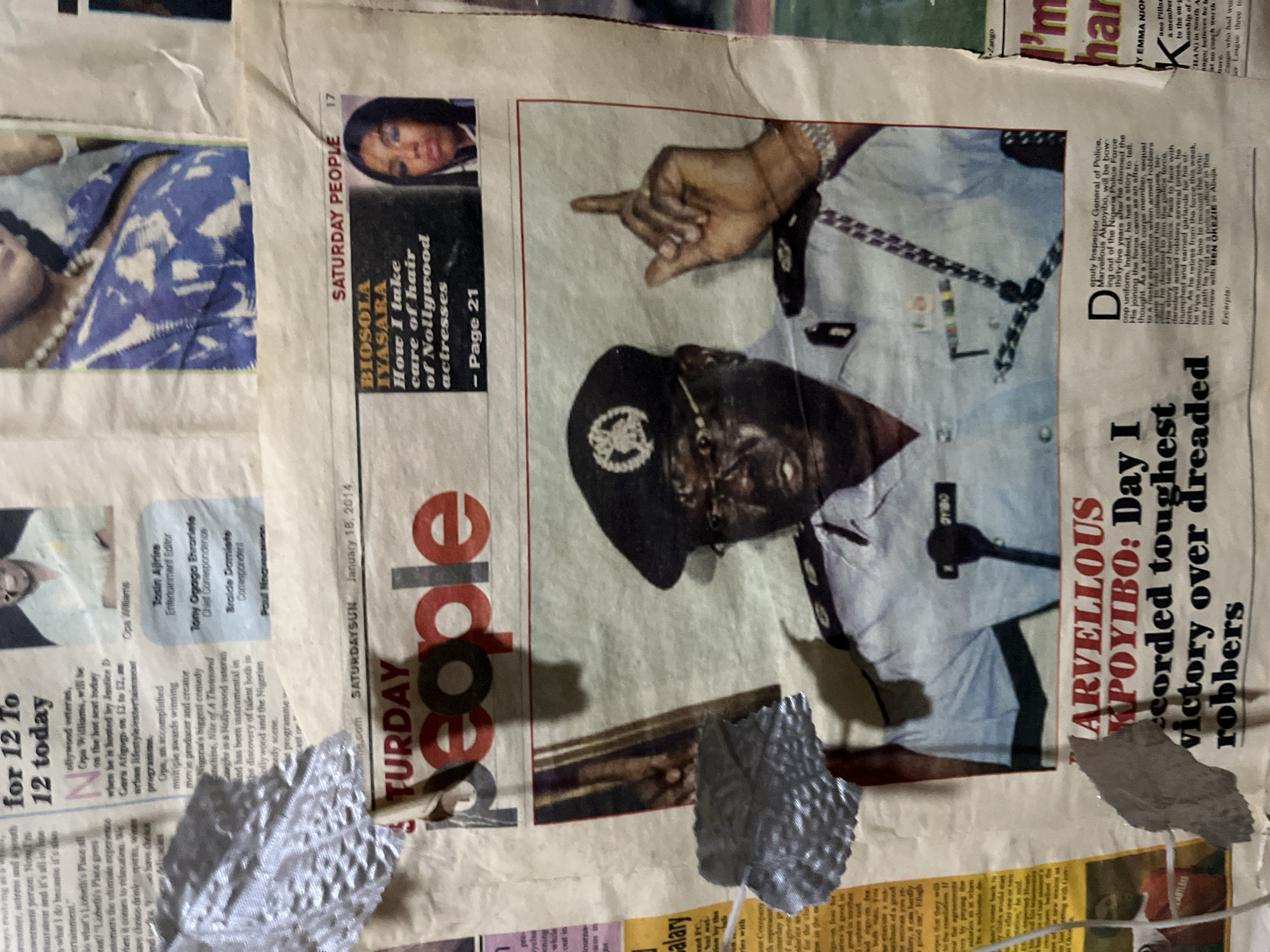 This is a close-up, slightly crumpled photograph of a newspaper page, likely displayed on a wall. The dominant image on the newspaper is a large photograph of a man in a Nigerian police uniform, identified in the headline as "MARVELLOUS AKPOYIBO: Day I recorded toughest victory over dreaded robbers." He is wearing glasses and a police cap with a badge, and has a serious expression. His right hand is raised, with his index finger pointing upwards.
To the upper right of this main image, there is a smaller inset photograph of a woman. Below this, a headline reads "BIOSOLA IYASARA How I take care of hair of Nollywood actresses - Page 21." Further up the page, a smaller article features a headshot of a smiling man with the name "Opa Williams" below it. Another smaller article mentions "Cos Williams."
The newspaper itself appears to be the "SATURDAY PEOPLE" edition, dated January 18, 2014. The overall image is lit from the front, casting shadows. The newspaper page is textured and shows signs of wear, with some crumpled and torn sections, and what appears to be silver duct tape adhering to it in places. The background is indistinct, but suggests a wall mounting. The location of the image is not explicitly stated within the visible content.