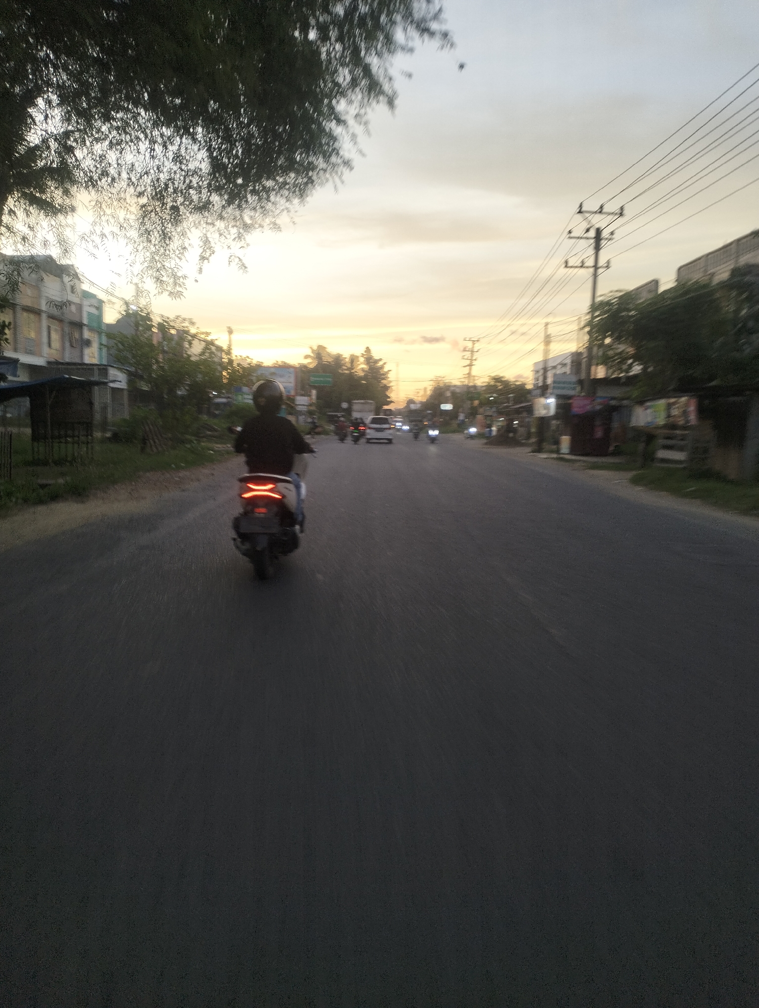This media file captures a scene on a road in Lhokseumawe, Indonesia, during what appears to be either sunrise or sunset. The sky is a gradient of soft oranges and yellows, transitioning to a pale blue, indicating dusk or dawn. A person on a scooter is the main subject, seen from behind, riding on the right side of the asphalt road. They are wearing a dark helmet and a dark top. The scooter's taillights are illuminated with a distinct red glow.
The setting is a semi-urban environment. On the left side of the road, there are buildings with visible signage, some appearing to be shops or small businesses, and a rudimentary shelter with a dark roof. Trees with dark foliage are present, with some branches extending into the frame from the top left, their leaves blurred due to motion. Power lines and utility poles are visible along the right side of the road, stretching into the distance, suggesting a developed area.
In the background, other vehicles, including at least one car and another scooter, are visible further down the road, also in motion, contributing to the sense of activity. The overall mood is calm and perhaps contemplative, possibly representing a commute or a leisurely ride as the day begins or ends. The image is slightly blurry, indicating movement, likely from the perspective of someone also traveling on a vehicle. No discernible text is clearly readable in the image.