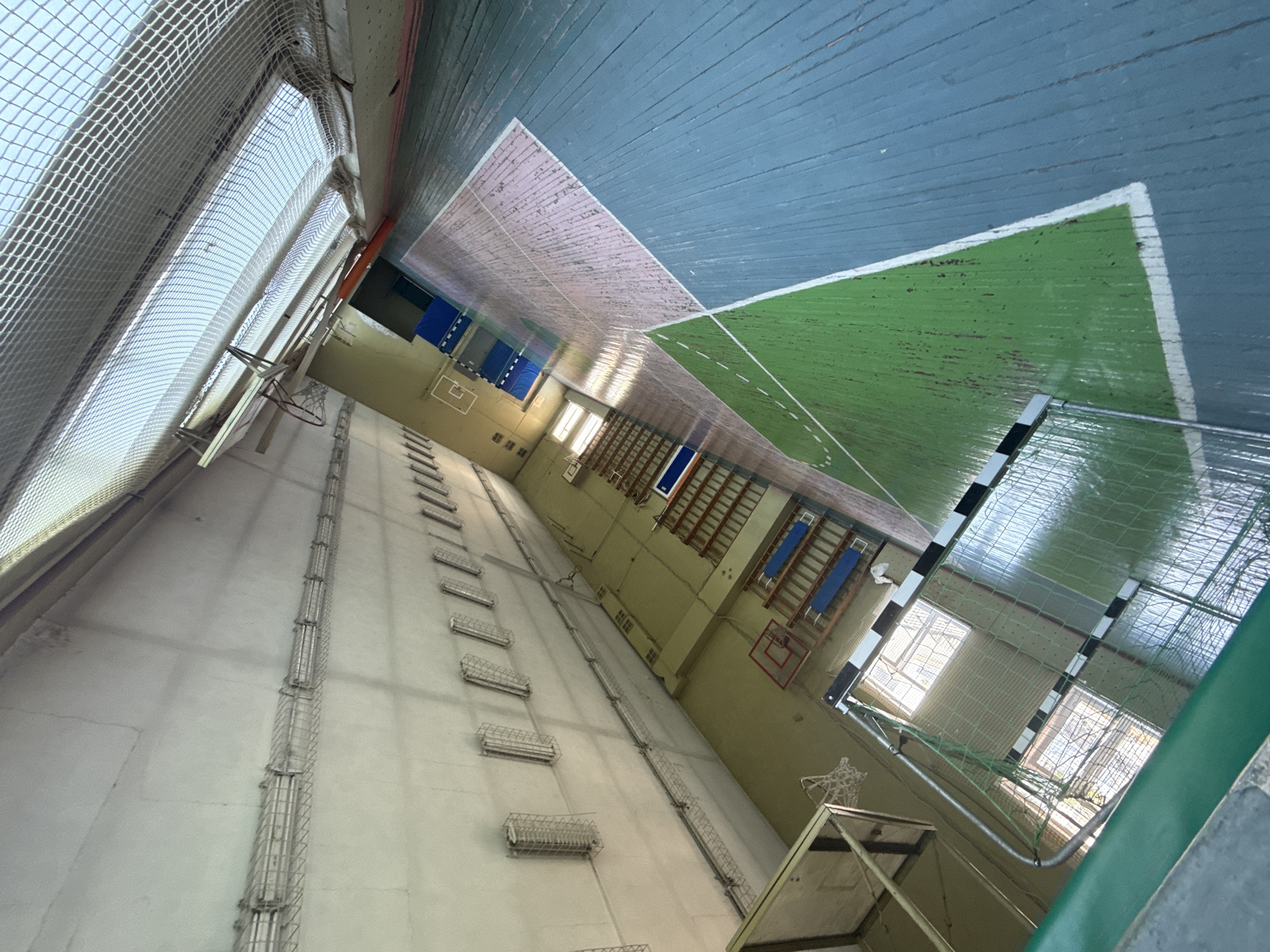 The image displays the interior of a brightly lit gymnasium, likely situated in Saint Petersburg, Russia. No people are present, and the sports hall appears ready for use. The floor is constructed from blue-painted wooden planks, featuring various white court markings. A distinct green rectangular playing area, outlined with white lines including a central dashed line, occupies a significant portion of the floor, suggesting a court for sports such as futsal or handball. At one end of this green court, a goal with characteristic black and white striped posts and a net is visible.
Along one of the longer walls, a series of wooden wall bars and climbing ladders are installed. Further along this wall and on what appears to be a back wall, basketball backboards with red-rimmed hoops are mounted. Large windows, providing ample natural light, are protected by a fine mesh netting, and similar protective netting extends across sections of the ceiling and upper walls. Elongated fluorescent light fixtures, enclosed within wire cages, are mounted along the walls to provide artificial illumination. The walls are painted in a utilitarian pale green or beige. The wooden floor shows noticeable wear and tear from extensive use. There is no visible text in the image.
