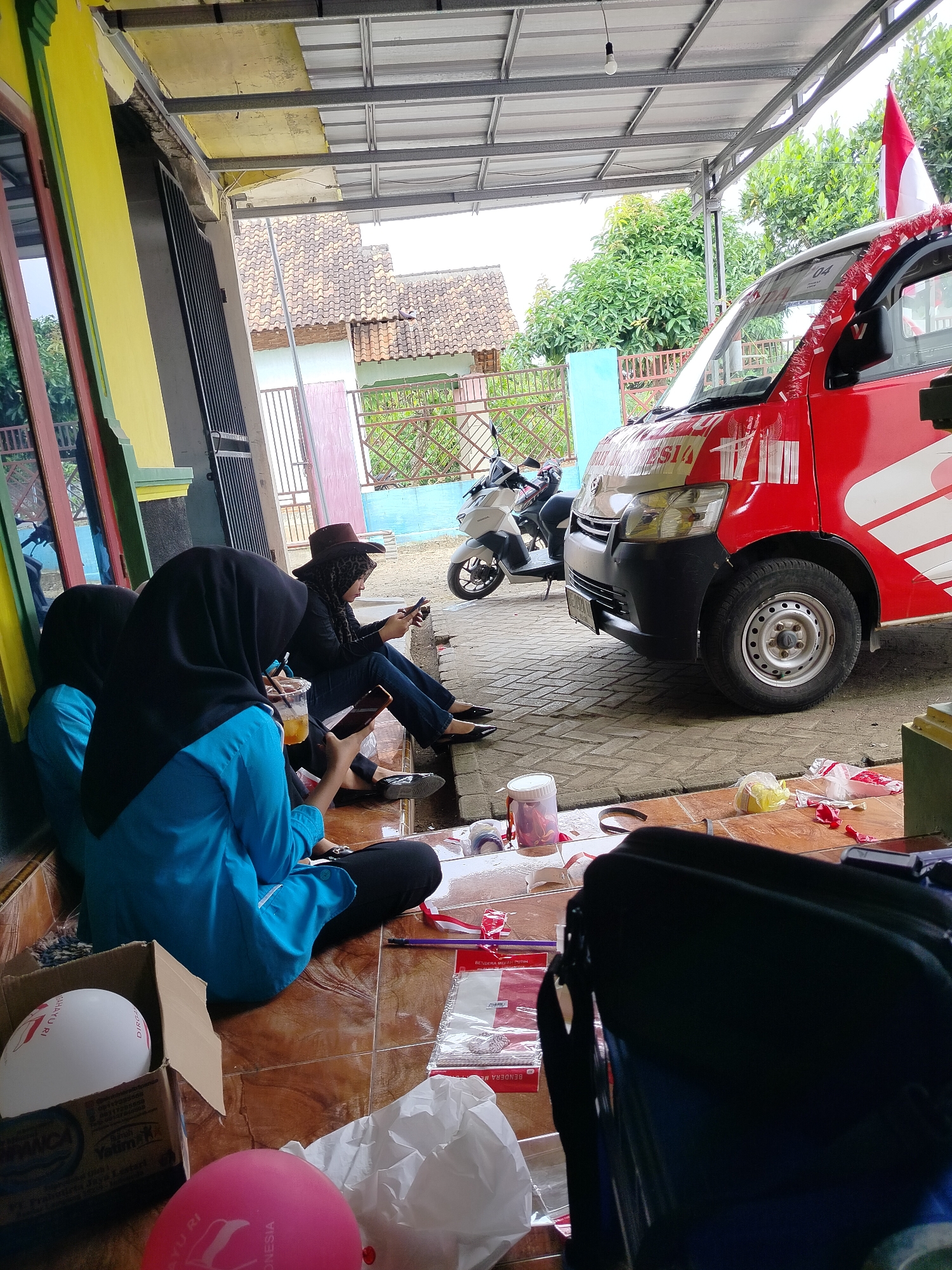 The image depicts a street-level scene in what appears to be a sheltered outdoor area, possibly a veranda or carport, in Sumberrejo, Indonesia. A red and white van, likely a delivery vehicle, is parked prominently on the right side of the frame. The van is decorated with red and white streamers and a small Indonesian flag is visible. In the foreground, three individuals are seated on the ground. Two individuals, dressed in blue tops and black hijabs, are looking at their phones. The third individual, wearing a brown hat and a black top, is also engrossed in their phone. Scattered on the tiled floor are various items, including a box with white and pink balloons, plastic cups, and some packaging materials. The background shows a residential building with tiled roofs and greenery. The overall atmosphere suggests a casual gathering or a break in activity.