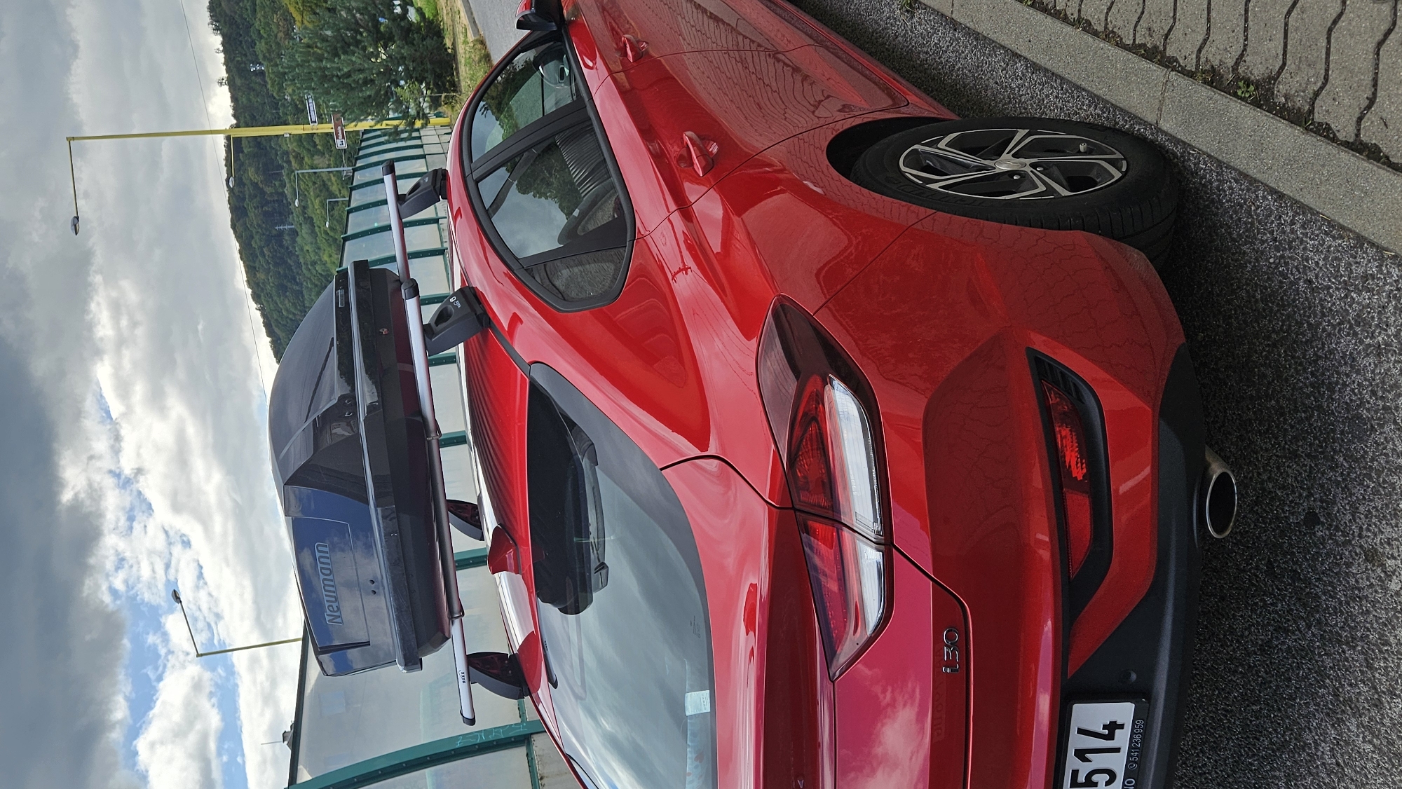 The image shows a bright red Hyundai i30 with a black roof box parked on a paved surface next to a curb. The car is facing away from the viewer, with its rear in the foreground. The roof box has the brand name "Neumann" written on it in blue letters. The background features a green, translucent barrier, a yellow lamppost, a forested hill, and a cloudy sky. The license plate on the rear of the car is partially visible, showing "514" and some Slovakian text. The overall scene suggests the car is parked on a street or a roadside area, possibly in a suburban or a less urbanized part of a city. The location of Prešov, Slovakia, is mentioned, implying this scene is within that city.