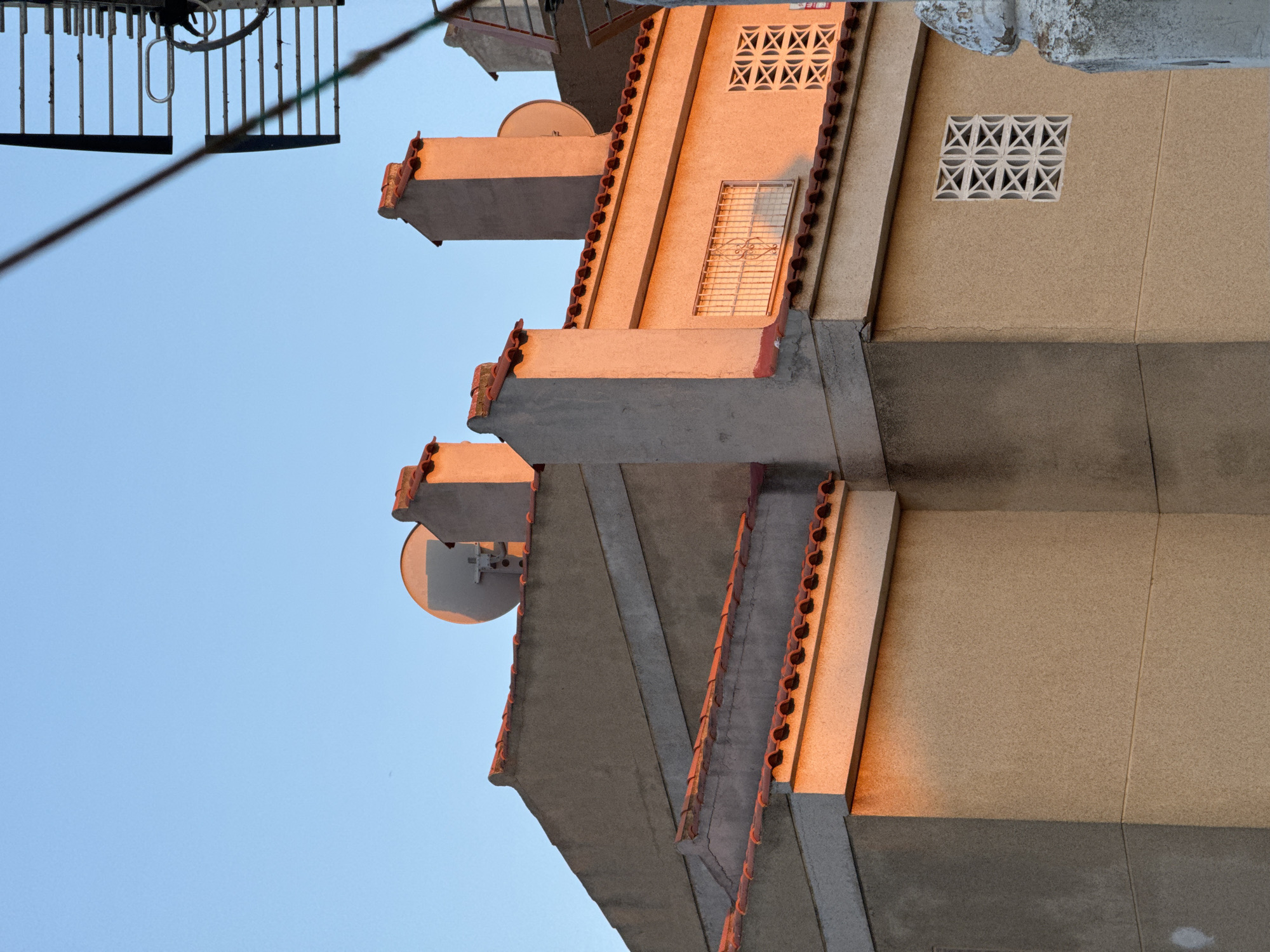 The image shows the side of a building in Torrevieja, Spain, at sunset. The building has a textured, light brown facade and a red tile roof. There are several chimneys on the roof, two of which are prominent in the foreground. One of the chimneys is illuminated by the warm glow of the setting sun. A satellite dish is visible on the roof behind the chimneys. The sky is a clear, pale blue, indicating good weather. In the upper right corner, a portion of a metal antenna structure is visible, partially obscuring the sky. A decorative white lattice pattern is visible on a section of the building's facade, next to a window with a metal grate. The overall impression is one of an urban residential area bathed in the golden light of dusk.