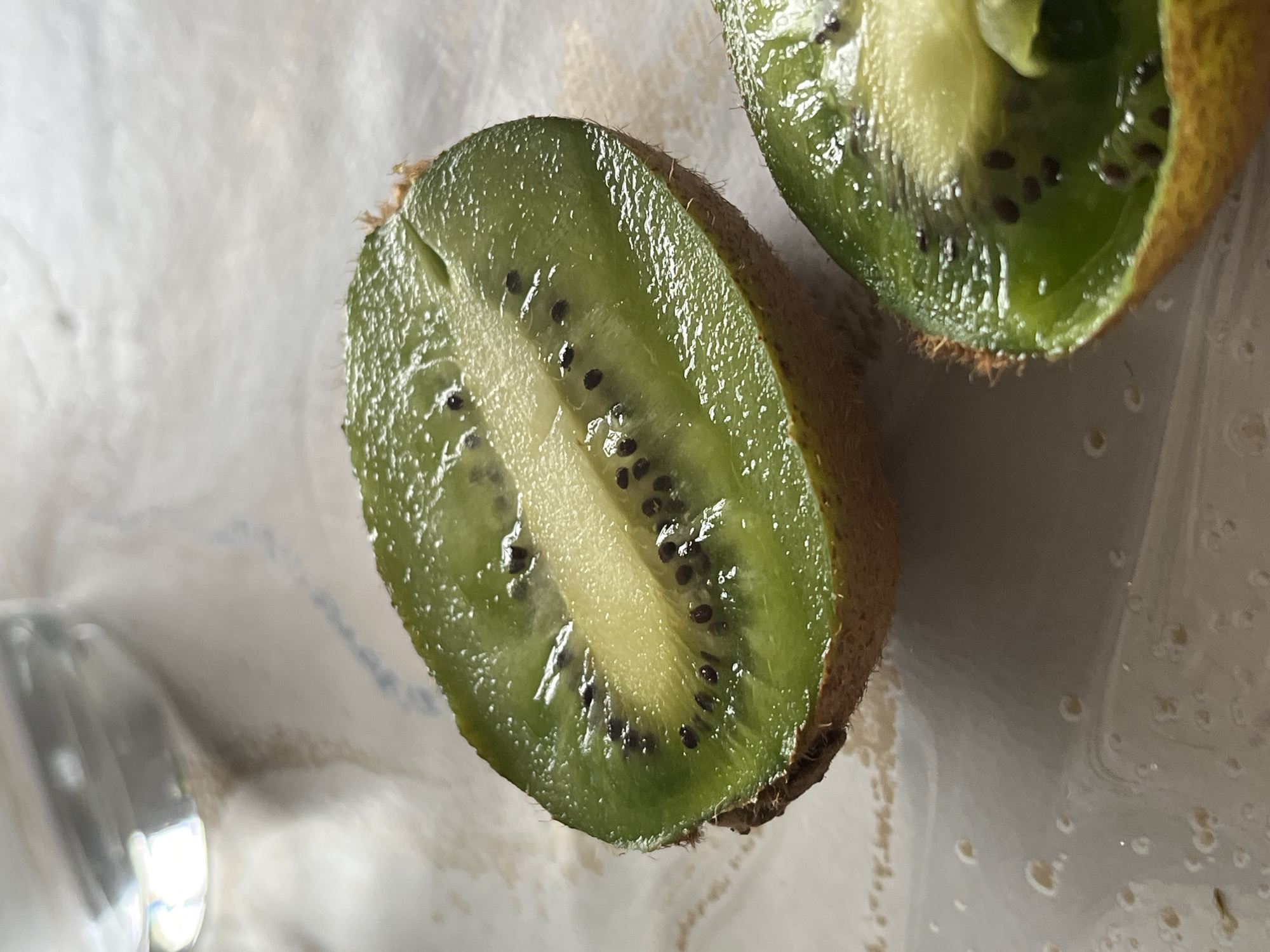 A close-up shot shows two halves of a sliced kiwi fruit resting on what appears to be a white surface with subtle patterns. The larger kiwi half is in sharp focus, revealing the green flesh speckled with tiny black seeds, surrounding a light yellow core. The fuzzy brown skin is visible around the edges of the fruit. The second kiwi half is slightly out of focus, positioned to the upper right of the first. The scene is brightly lit, highlighting the glossy texture of the kiwi.
There is no indication of the location being Saarbrücken, Germany, nor of any people or actions taking place. The focus is solely on the two kiwi halves and the surface they rest on.