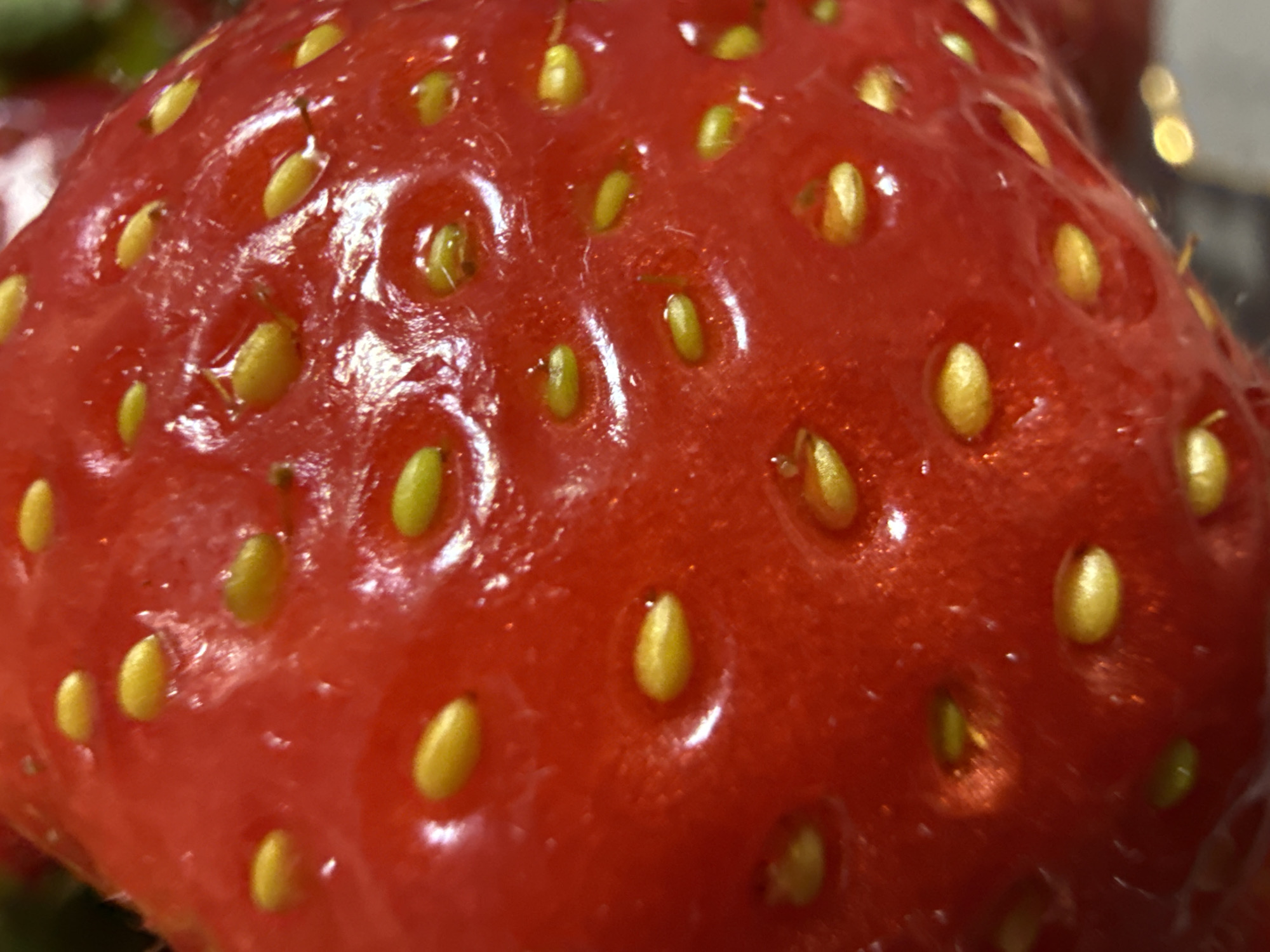 A close-up photograph shows the surface of a strawberry. The strawberry is red and glossy, with small, yellow seeds embedded in its surface. The surface is slightly uneven, with small indentations around each seed. The lighting creates highlights and shadows, emphasizing the texture of the fruit. The composition focuses entirely on the strawberry's surface. There is no location.