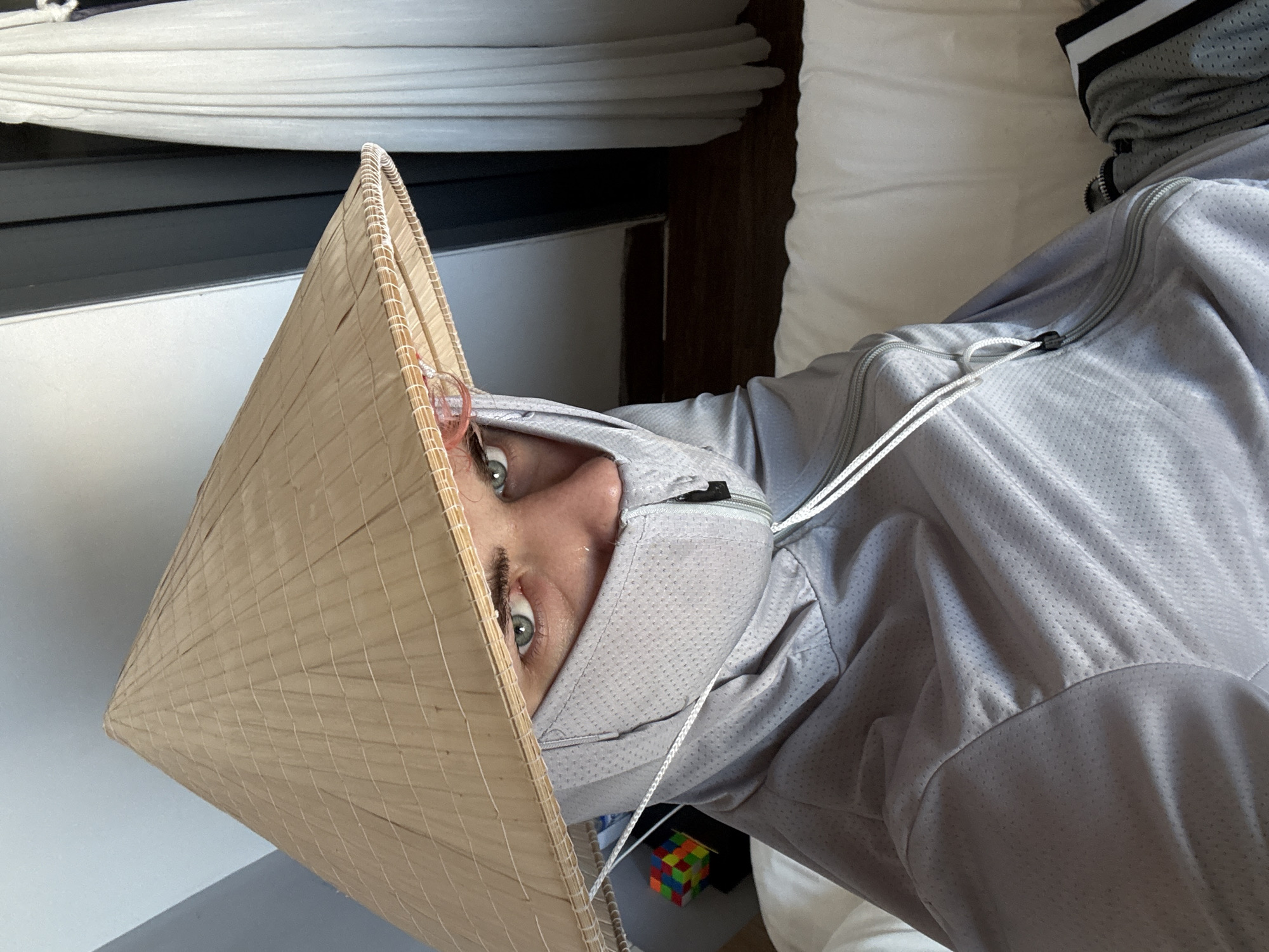 A person wearing a gray hooded jacket and a Vietnamese conical hat is looking directly at the camera. The person's face is partially obscured by the hat and the hood. Only one eye is fully visible. The background includes a window with drawn blinds and a white pillow. A Rubik's cube is partially visible near the bottom of the frame. The location is not identifiable from the image.