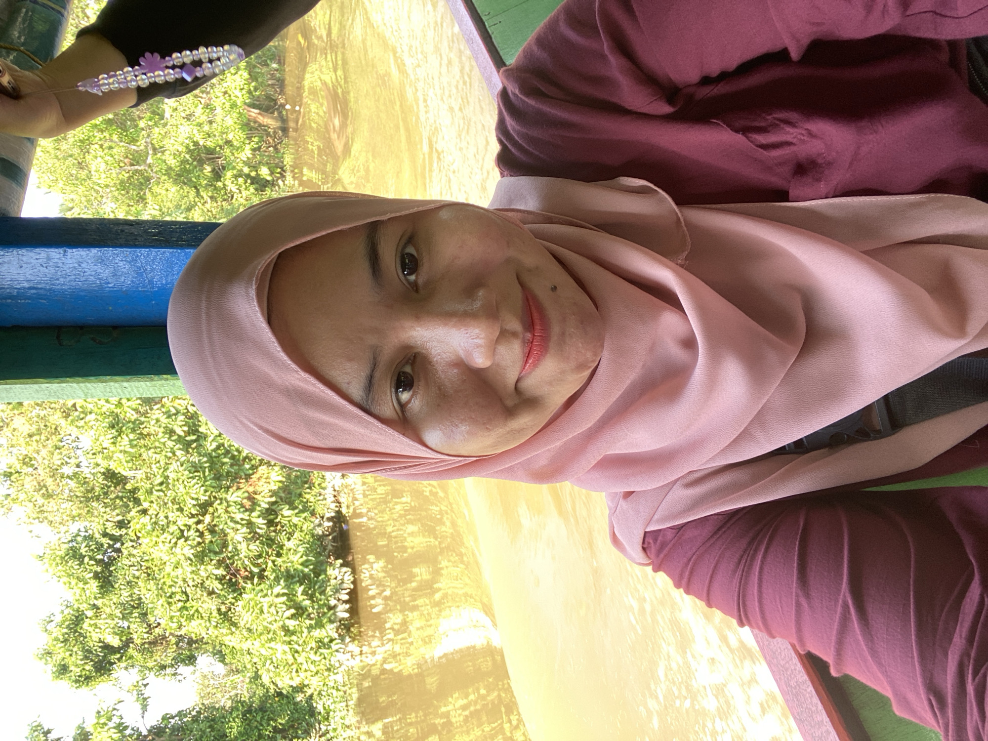 The image is a selfie taken by a woman wearing a pink hijab and a maroon long-sleeved shirt. She is on a boat, with the muddy, yellowish water of a river visible in the background. Lush green trees line the riverbanks, suggesting a natural, possibly rural, environment. The boat appears to be a traditional wooden vessel with green and blue painted sections along the railing. A beaded bracelet with purple charms is visible on another person's hand in the upper right corner of the image. The overall impression is of a casual outing on a waterway, characteristic of the riverine landscape of Banjarmasin, Indonesia, known for its extensive network of rivers and canals.