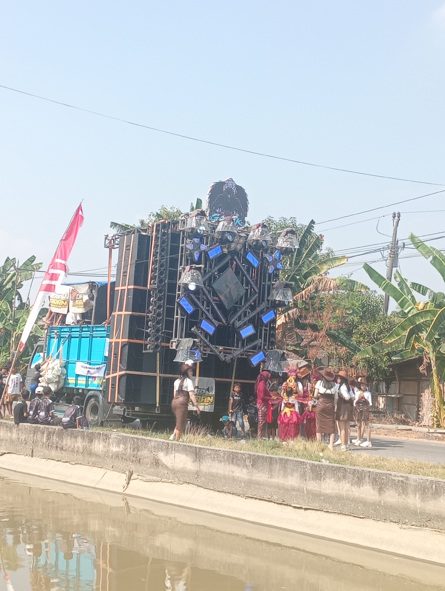 A group of people are gathered near a large sound system mounted on a truck. The sound system is elaborate, with many speakers stacked vertically and a large, dark structure with lights above it. A red and white flag is attached to a pole near the truck. In the foreground, a concrete canal runs horizontally, with water reflecting the scene. On the side of the road, a group of people in traditional attire, including women in brown skirts and white tops with hats, are standing. To the left, children and adults are seated and standing near the truck. The environment appears to be outdoors on a sunny day, with trees and a power pole visible in the background. The scene suggests a festive gathering or event.