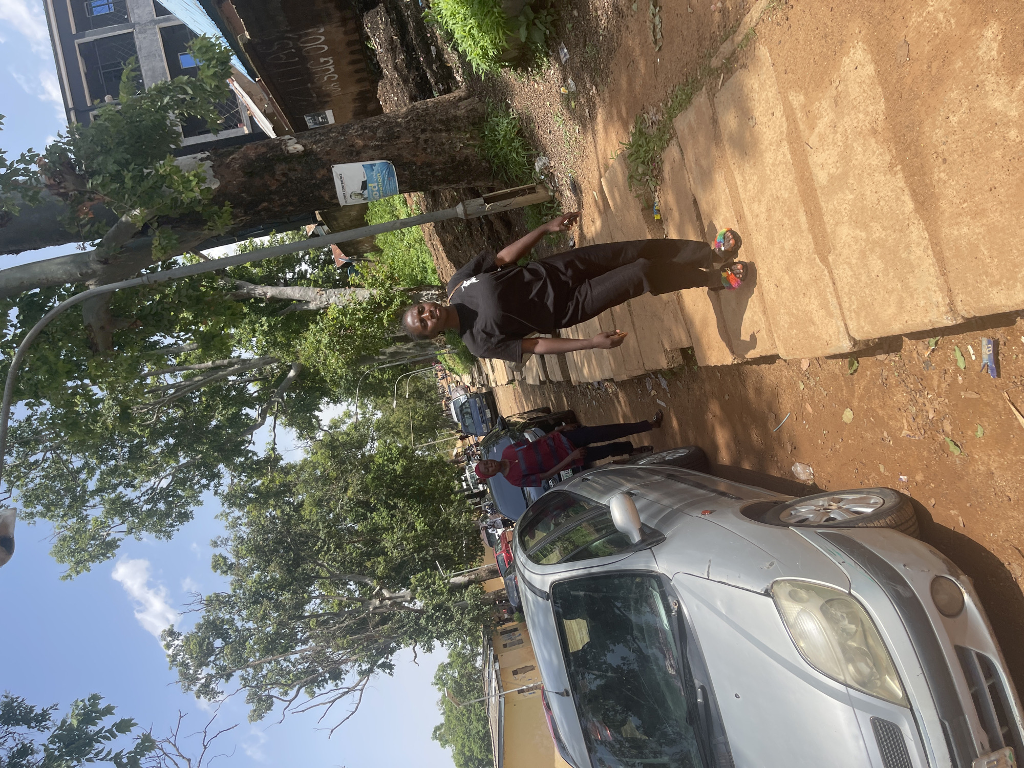 A low-angle, outdoor, eye-level shot captures a young person standing on a dirt path in what appears to be an urban setting in Jos, Nigeria. The person, wearing a black t-shirt, dark pants, and colorful slides, is posing for the camera. Their stance is casual, with one hand extended slightly outward. The scene is bathed in daylight, suggesting it is daytime.
Behind the individual, a large tree with lush green foliage dominates the left side of the frame, its trunk thick and weathered. A poster is affixed to the tree, but the text is partially obscured and illegible. Further back, a multi-story building with dark windows and balconies stands prominently. To the right of the tree and the person, a paved walkway is visible, leading to a dusty ground area with some scattered debris.
In the foreground, a silver car is parked, its front end slightly angled towards the viewer. The car appears to be a hatchback with a visible dent on its hood. Another car, partially visible, is parked behind it. The overall environment suggests a busy street or residential area, possibly with parked vehicles lining the side. The ground is a mix of dry dirt and concrete. The sky is visible in the background, a pale blue with scattered white clouds. The overall mood of the image is relaxed and everyday, capturing a moment in time on a sunny day in Nigeria.