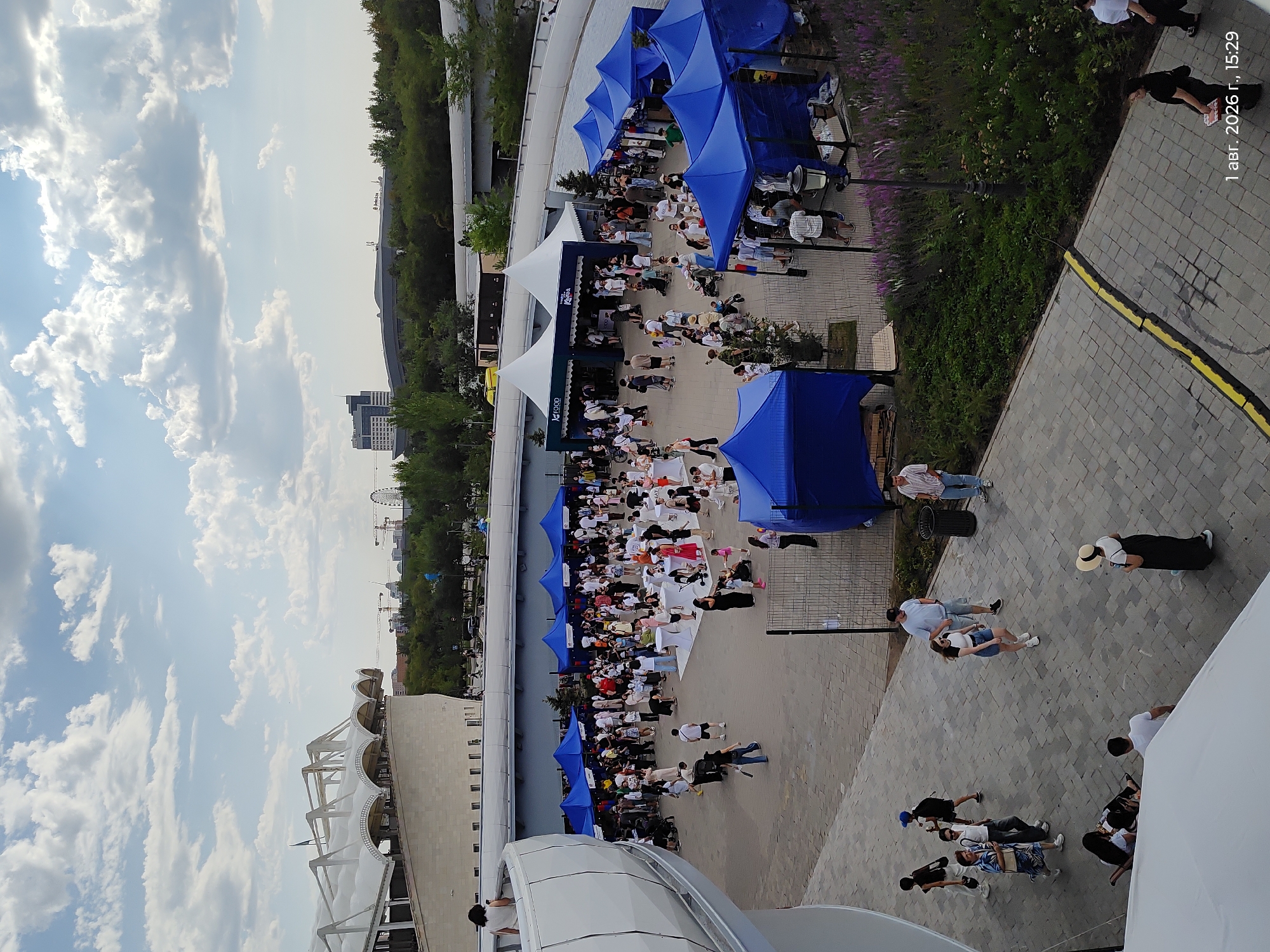 An aerial view captures a vibrant outdoor market or festival event taking place in Astana, Kazakhstan, likely near the "Nur Alem" Pavilion of the EXPO 2017 grounds, which is visible in the background. The scene is characterized by numerous blue and white tents, some of which appear to be food stalls, set up on a paved plaza. A large crowd of people, dressed in casual summer attire, are milling about, interacting, and browsing the stalls. Children are visible, some with strollers. The environment includes lush green trees and landscaping, a modern architectural structure with a white curved roof in the foreground left, and a cloudy sky. The lighting suggests it is daytime, possibly late afternoon due to the timestamp visible in the lower right corner of the image: "1 авг. 2026 г., 15:29" (August 1, 2026, 15:29). People are engaged in various activities: walking, standing in groups, and interacting with vendors. Some individuals are seated on the ground near the modern architectural element. The overall atmosphere appears festive and social.