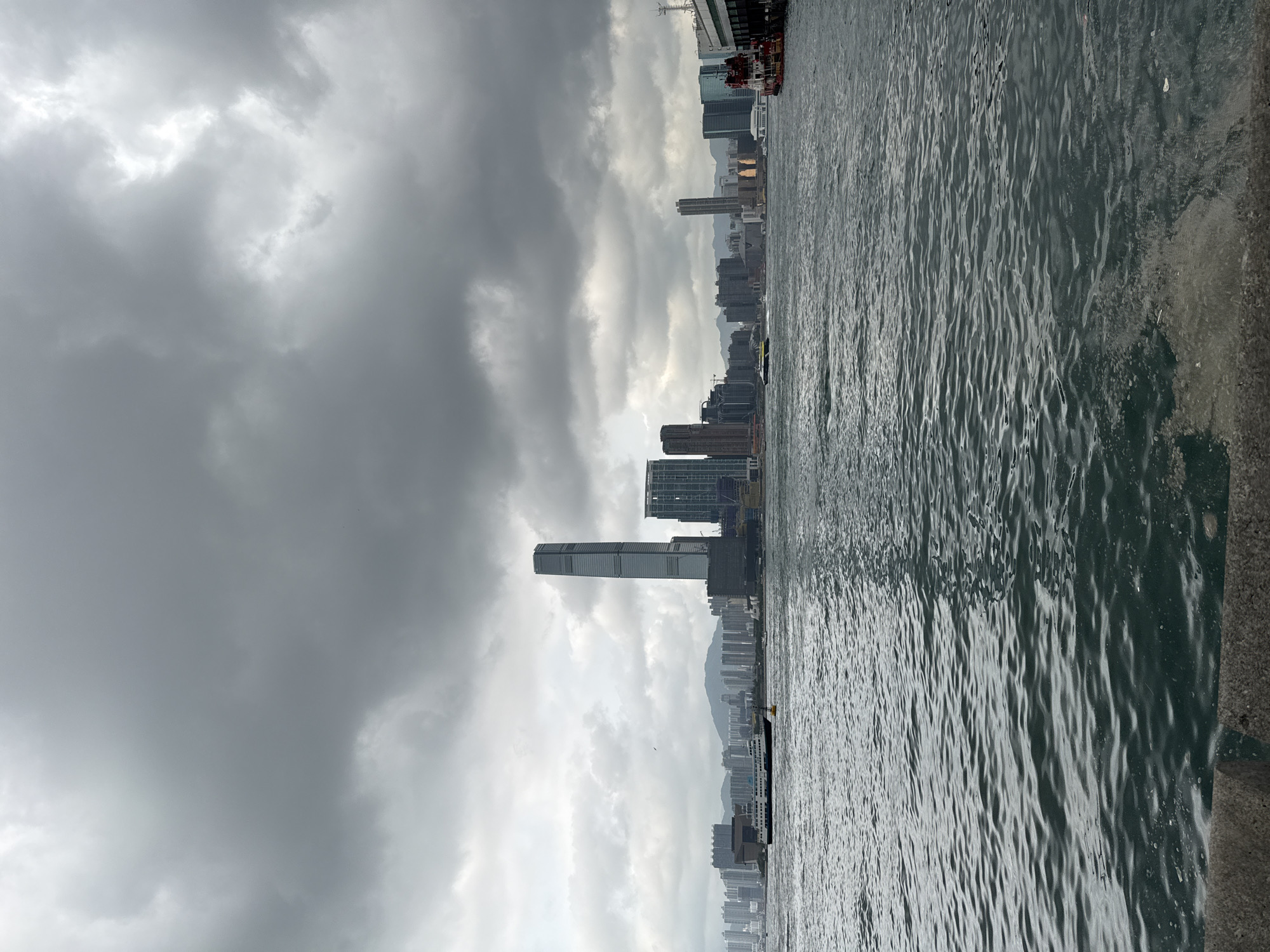 The image shows a cityscape under a cloudy sky, adjacent to a body of water. Tall buildings and skyscrapers are visible, typical of a dense urban environment. The water's surface is rippled, suggesting the presence of wind. The photograph appears to be taken from a low vantage point, close to the water's edge. The location is Hong Kong, China.