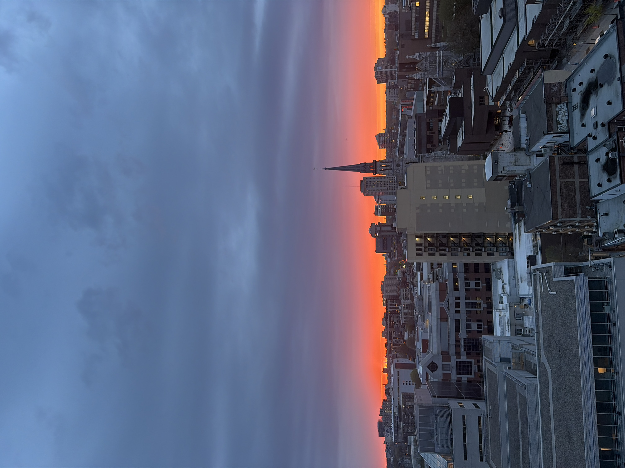 The image captures a sunset view of the Montreal skyline in Canada. The sky displays a gradient, transitioning from dark blue on the left to a vibrant orange at the horizon. A distinct layer of purple separates the blue and orange. The cityscape is silhouetted against the sunset, with a prominent church steeple rising above the other buildings. The buildings are a mix of high-rise and low-rise structures, with details like windows and rooftops visible. The picture is taken from an elevated perspective, likely from a rooftop or a high-rise building.