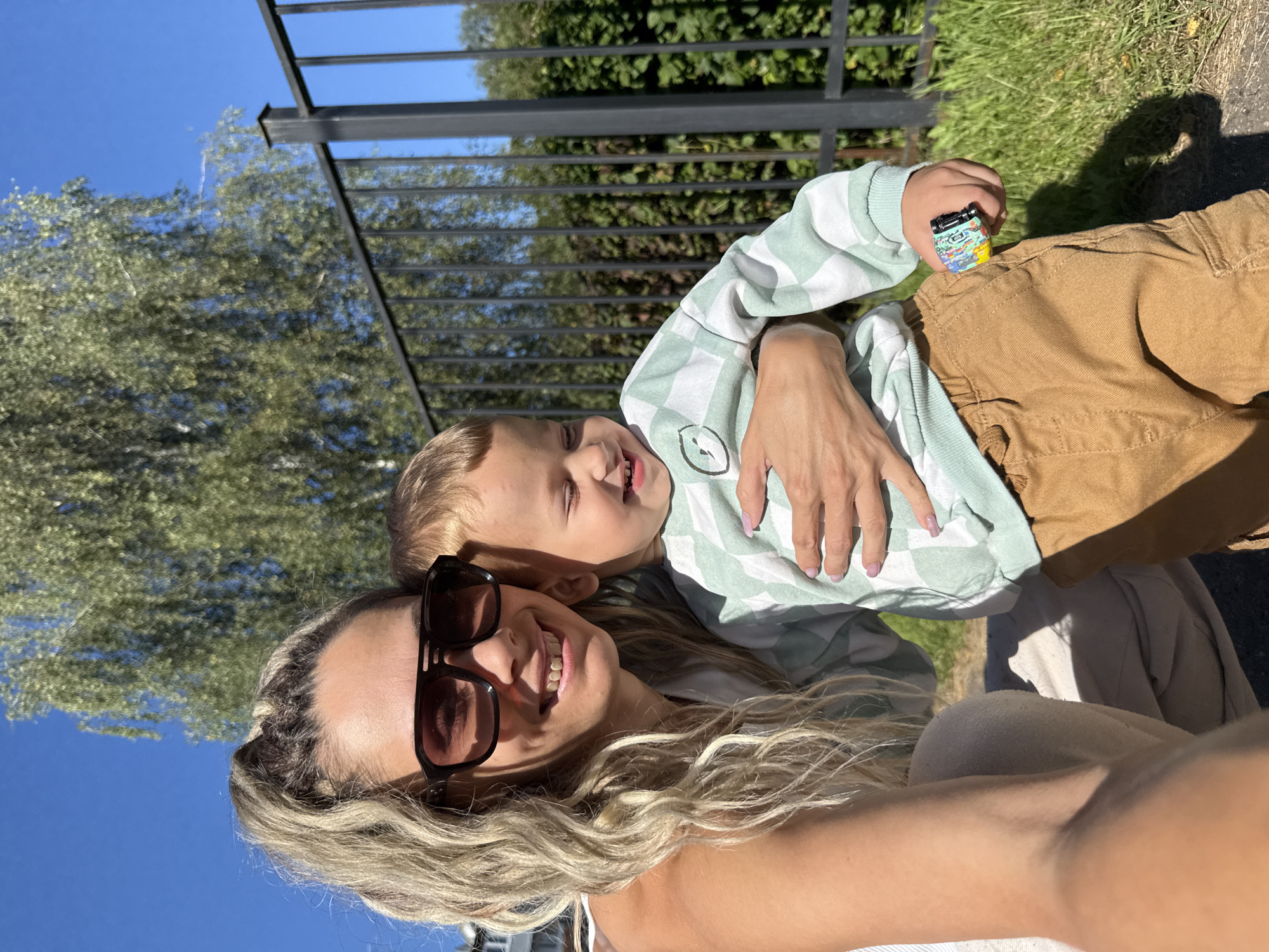 In Rusanovo, Russia, a woman with blonde hair, wearing large sunglasses, is holding a young child. The child is wearing a mint green and white checkered sweater and brown pants. The child is smiling and appears to be laughing. The woman is also smiling. They are standing outside, with a black metal fence and green foliage in the background. A large tree is visible above the fence, and the sky is clear and blue. The child is holding a small, colorful toy in their right hand. The lighting suggests it is a sunny day.