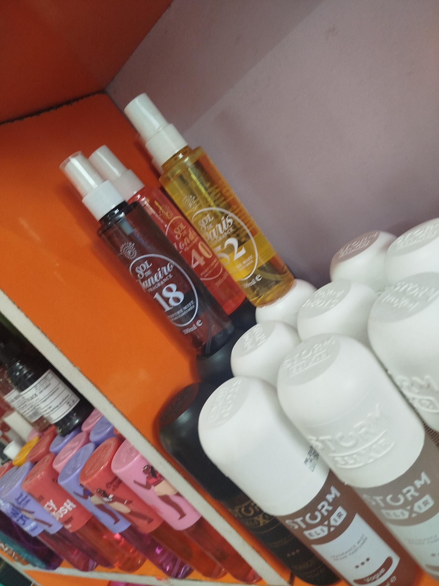 The image depicts a shelf in a store located in Ugbowo, Nigeria, displaying various cosmetic and fragrance products. On the top shelf, three spray bottles are visible: one with a clear yellow liquid and the brand name "SOL de Janeiro" along with the number "40", a dark brown bottle also with "SOL de Janeiro" branding and the number "18", and a partially visible red bottle. Below these, a row of smaller, brightly colored bottles with patterned caps are arranged, including purple, pink, and red containers with cartoonish figures printed on them. Further to the right, a cluster of white, cylindrical containers with embossed branding are stacked. The shelves are made of wood and painted orange and white, suggesting a retail environment. The lighting is dim, casting shadows on the products.