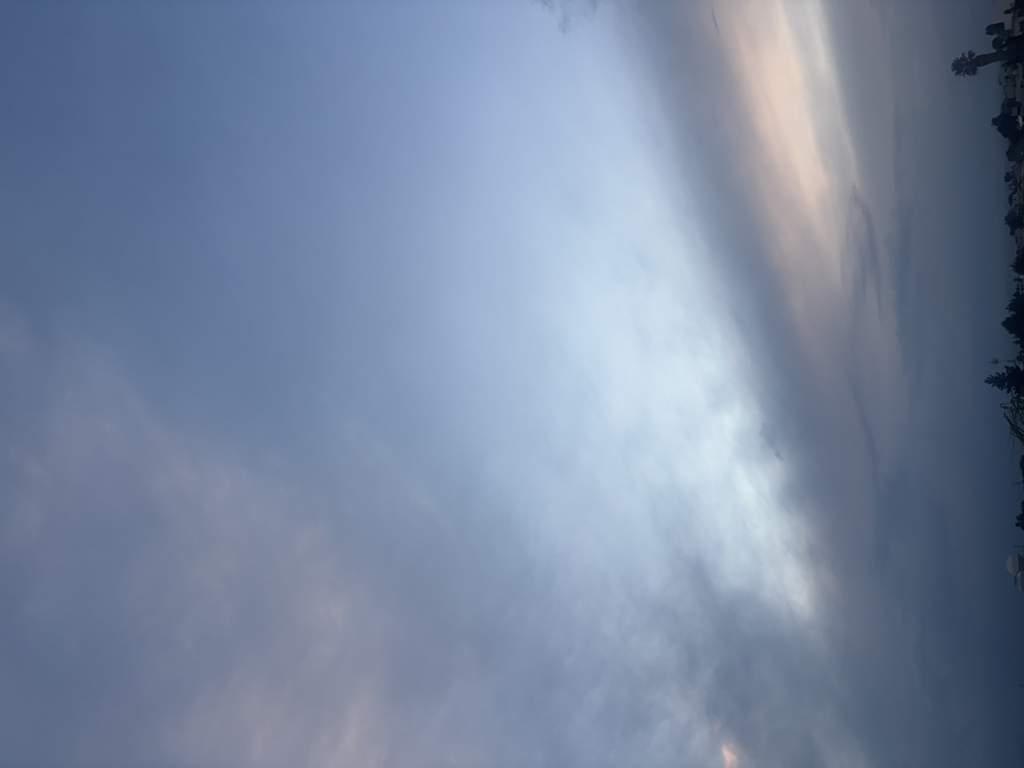 The image shows a view of the sky in San Pablo del Monte, Mexico. The sky is predominantly blue with variations in tone and wisps of clouds. A darker area appears to be approaching from the right side of the frame, with what seems to be a layer of darker clouds. Along the very right edge, silhouetted trees, and a glimpse of urban structure are visible, suggesting the location within the city. The lighting seems to indicate either sunrise or sunset, given the color variations in the clouds. No people, specific objects, actions, or interactions are visible in the sky itself. The environment is focused on the atmospheric conditions.