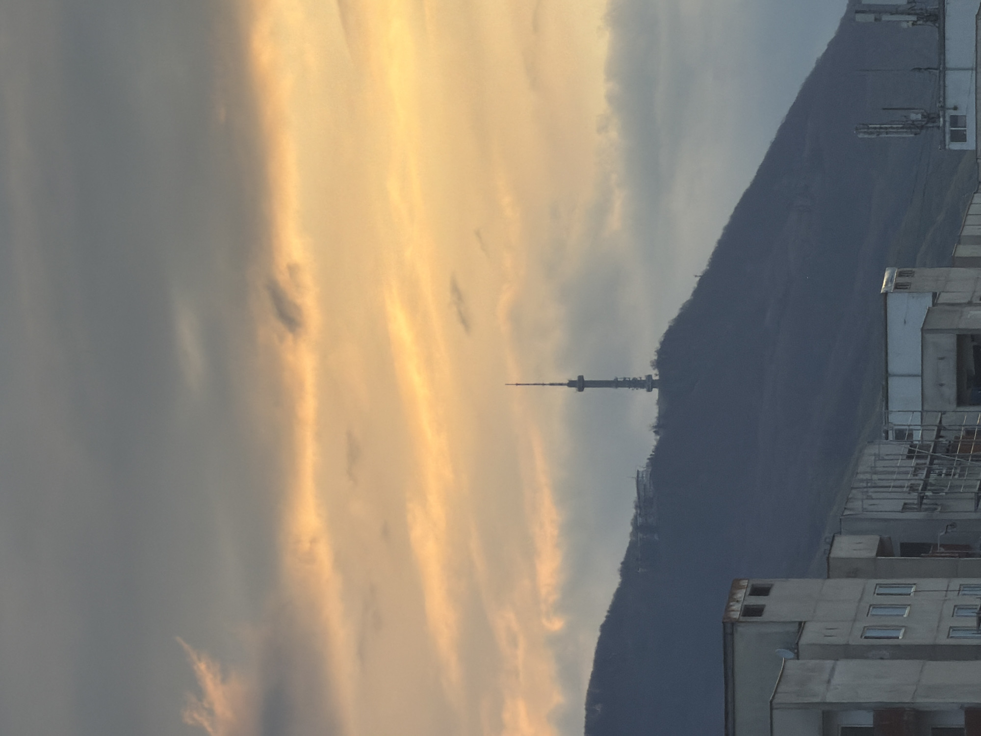 The image, correctly oriented, presents a panoramic view of an urban landscape backed by a mountain under a dramatic sky, characteristic of either sunrise or sunset. The sky occupies the upper portion, displaying a striking gradient from dark grey, heavily clouded areas on the left to vibrant golden and orange clouds brightly illuminated by the sun on the right. These clouds suggest a partly cloudy day during the "golden hour."
Dominating the midground is a large, dark mountain, its slopes appearing densely forested, stretching horizontally across the frame. Perched distinctly on a peak or high ridge of this mountain, near the center, is a tall communication tower with a visible antenna structure. This is highly indicative of the Kopitoto TV Tower on Vitosha mountain, given the location context of Sofia, Bulgaria.
In the immediate foreground, on the right side of the image, are parts of several multi-story buildings, likely residential or commercial structures. They appear to be constructed from light-colored concrete, with visible windows. Some rooftops feature what appear to be utility installations or additional antenna arrays.
No people or explicit activities are visible, presenting a static scene. There is no discernable text within the image.