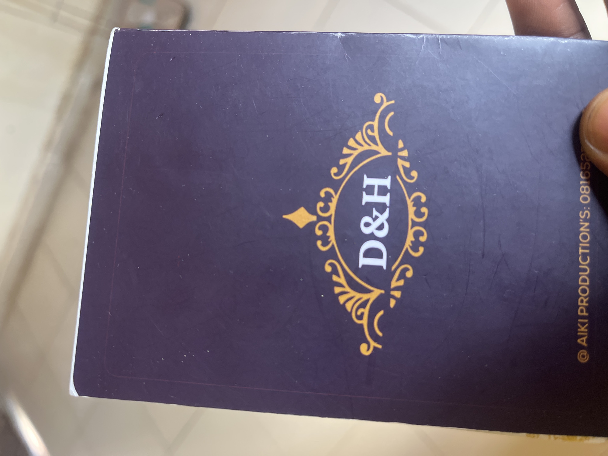 A close-up, low-angle shot shows the cover of a business card with a dark purple background. In the center, the initials "D&H" are displayed in white, bold, sans-serif font, enclosed within an ornate gold emblem that features swirling decorative elements and a diamond-shaped accent above the initials. At the bottom of the card, the text "@ AIKI PRODUCTION'S: 081652..." is visible in white. The card appears to be held by a person's fingers, partially visible at the bottom right. The background suggests an indoor setting with a blurred, tiled surface and a wall. The image does not provide specific information to locate it within the city of Jalingo, Nigeria.