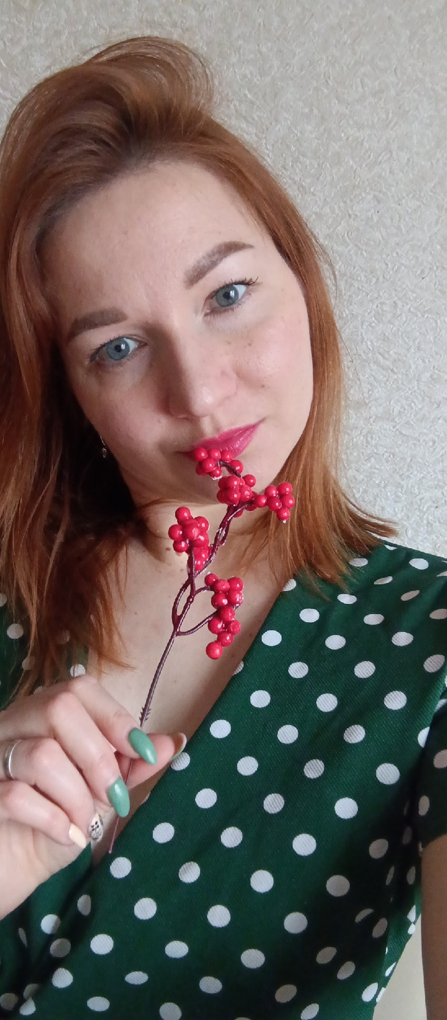 A woman with auburn hair and blue eyes is holding a sprig of red berries near her face. She is wearing a green dress with white polka dots and has light green nail polish. A silver ring is visible on her left ring finger. The background is a textured, light-colored wall. The location and city are unknown.