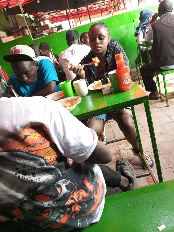 The scene takes place in an outdoor eatery with green tables and benches, located in Machakos, Kenya at coordinates -1.506 latitude, 37.266 longitude. A young woman wearing a black jacket and jeans is sitting at a table, eating from a plate and holding a fork. She is wearing a red and white necklace. There is a juice bottle, two plastic cups, and a salt shaker on the table. A young man wearing a blue shirt and a white cap sits at the table next to her. He is looking down. A man with a white shirt sits to his left, holding a white bag. A person wearing a white shirt with an orange and black skull design is sitting at a green bench to the left of the woman. An older man wearing a brown jacket, blue jeans, and a white shirt sits at a separate table. He is looking down and speaking to a younger man wearing a blue shirt and a black cap. Behind the man in the brown jacket is a younger man wearing a blue shirt and a white cap.
