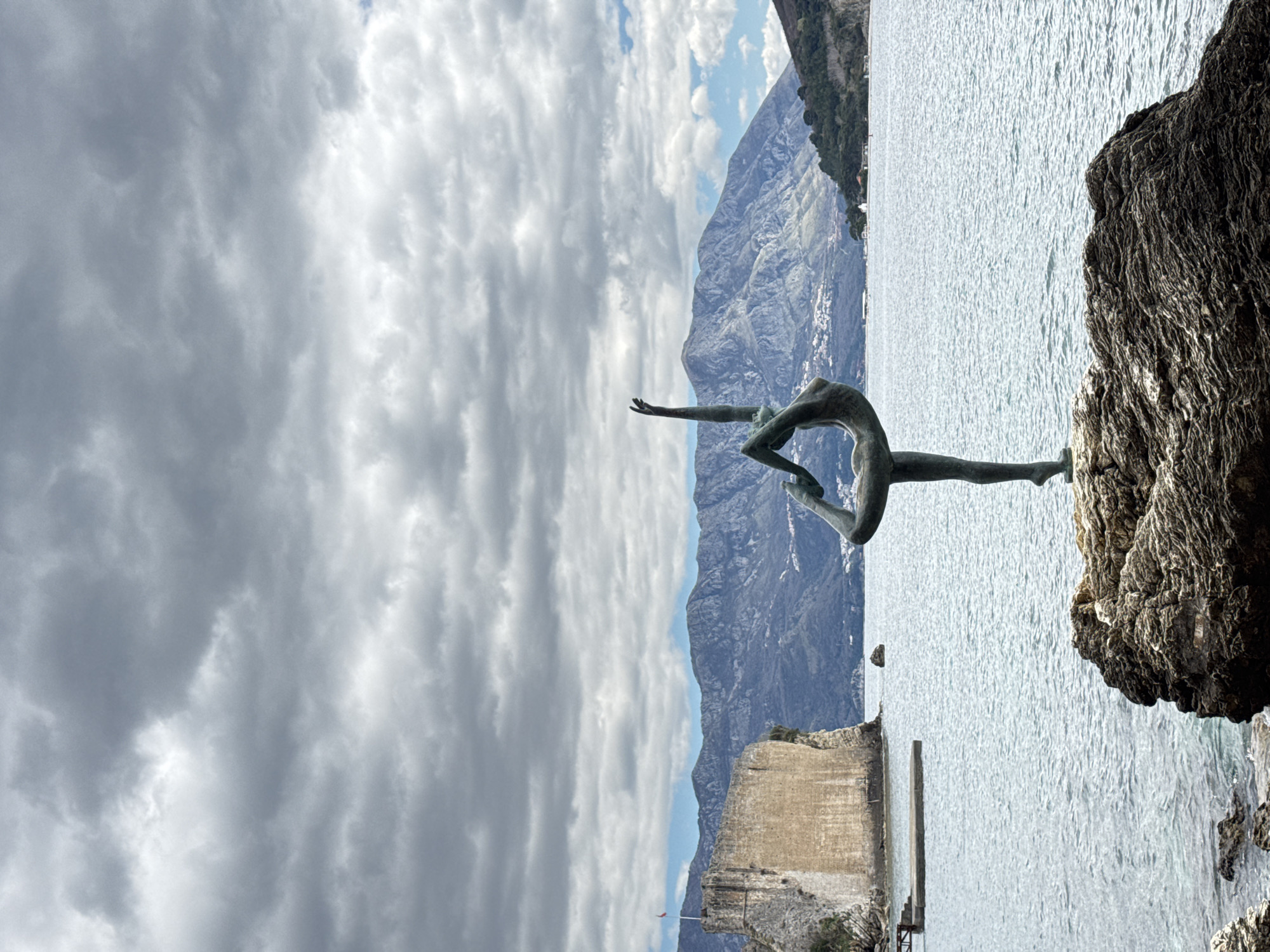 The image is of the “Ballerina of Budva” statue in Budva, Montenegro. The statue is located on a rock overlooking the Adriatic Sea with mountains in the background. There is a cloudy sky. The statue is of a woman in a contorted yoga position. To the left of the statue, there is a stone ruin jutting out into the water.