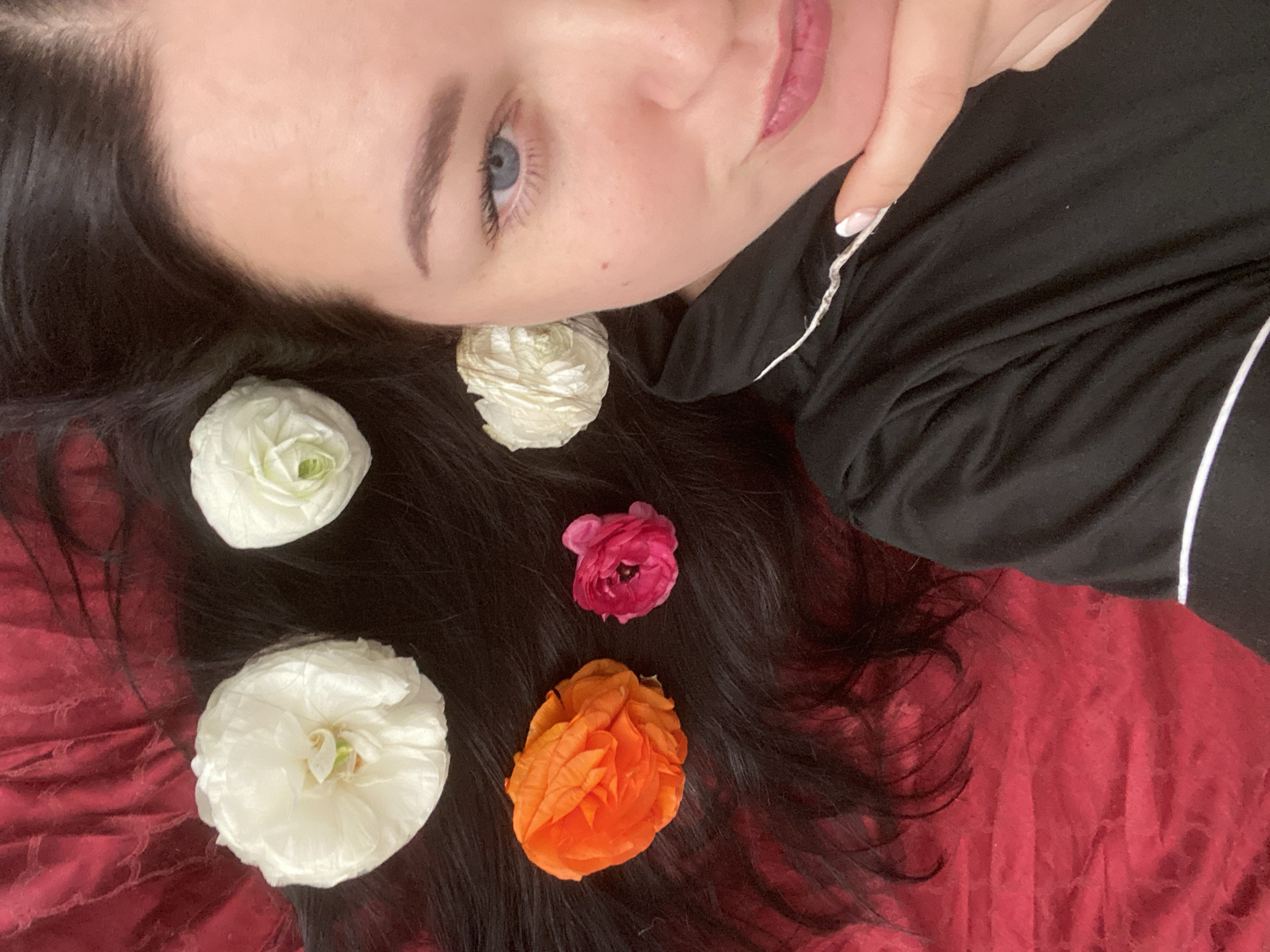 The image captures a woman lying down, viewed from an overhead perspective, with her head and shoulders filling the frame. She has dark, long hair spread out against a crumpled, deep red fabric, which appears to be a bedspread or blanket. Her face, characterized by fair skin and serene blue eyes, is angled slightly towards the viewer. Her eyebrows are well-groomed, and her lips, adorned with a glossy pink shade, are slightly parted.
She is wearing a black top or pajama shirt with white piping detail. Her right hand is gently positioned near her chin, revealing manicured fingernails with a French tip design. Artistically arranged within her dark hair are five ranunculus flowers: two white, one off-white/cream, one vibrant orange, and one fuchsia pink. The overall scene suggests a calm and contemplative mood. The lighting is soft, indicating an indoor setting. No specific location cues are visible within the image, but the provided context is Sochi, Russia. There is no visible text in the media file.