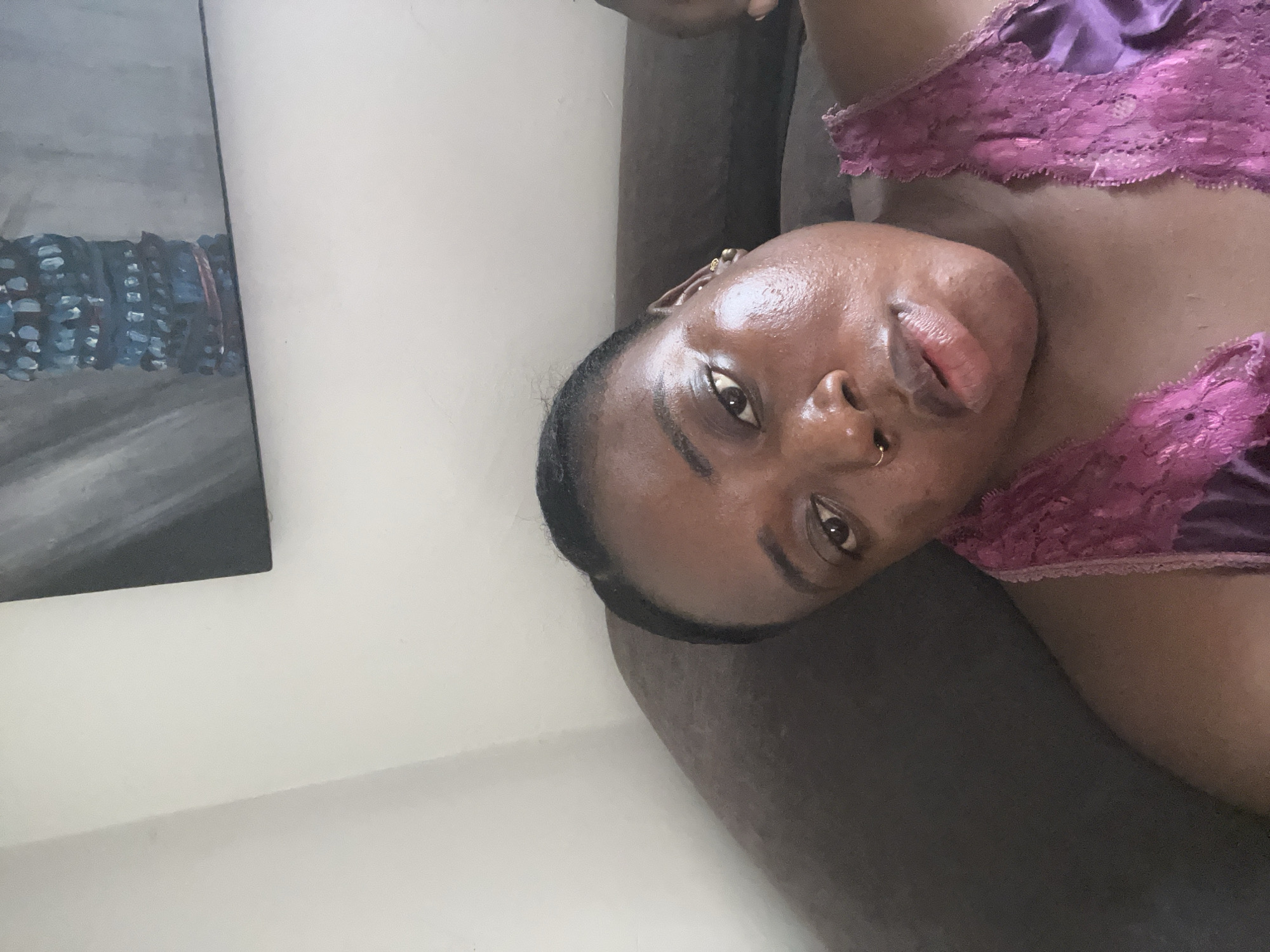 A dark-skinned woman wearing a purple lacy camisole lies on a brown sofa. A piece of art hangs on the white wall behind the sofa. It is difficult to determine the precise location in Abuja, Nigeria, without further information.
