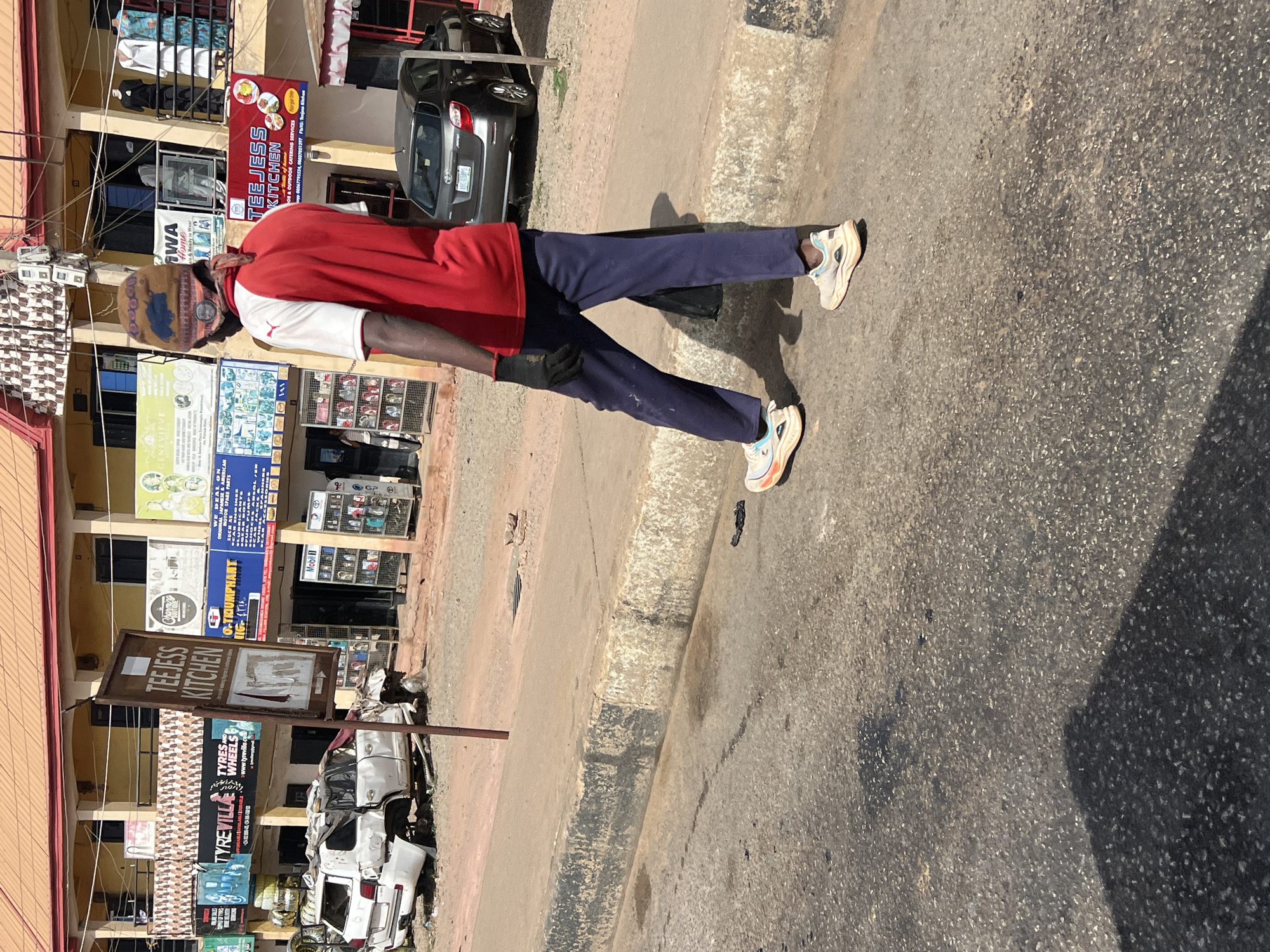 The image is taken outdoors on a sunny day in Gura Top, Nigeria, in a commercial area with buildings advertising businesses. The main subject is a man wearing a red and white Puma jacket, dark blue pants, black gloves, and sneakers. He has a patterned hat or head covering and a face mask. He is walking on the pavement.
The buildings in the background feature a variety of signs. Some of the businesses that are visible include "Teejess Kitchen," "Tyrevilla," and "O-Triumphant Orion" selling motor spare parts. A wrecked white vehicle sits near the base of a sign for "Teejess Kitchen." Other cars are parked nearby. The street appears to be asphalt.