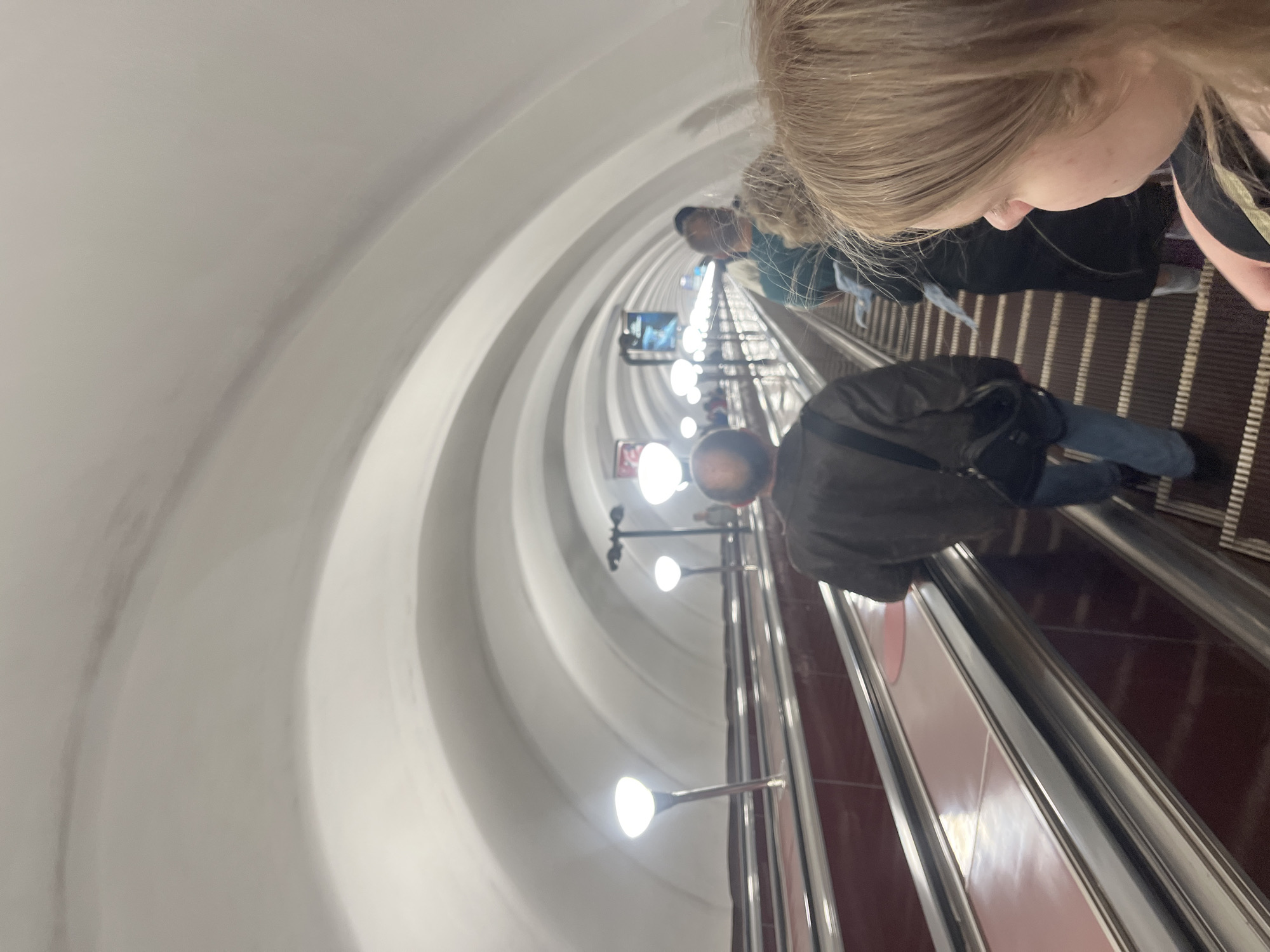 The image shows the interior of a metro station in Saint Petersburg, Russia. An escalator is prominently featured, descending into the station. People are riding the escalator. The architecture of the station includes a curved ceiling with evenly spaced light fixtures. The escalator steps are brown. The escalator rails are chrome.