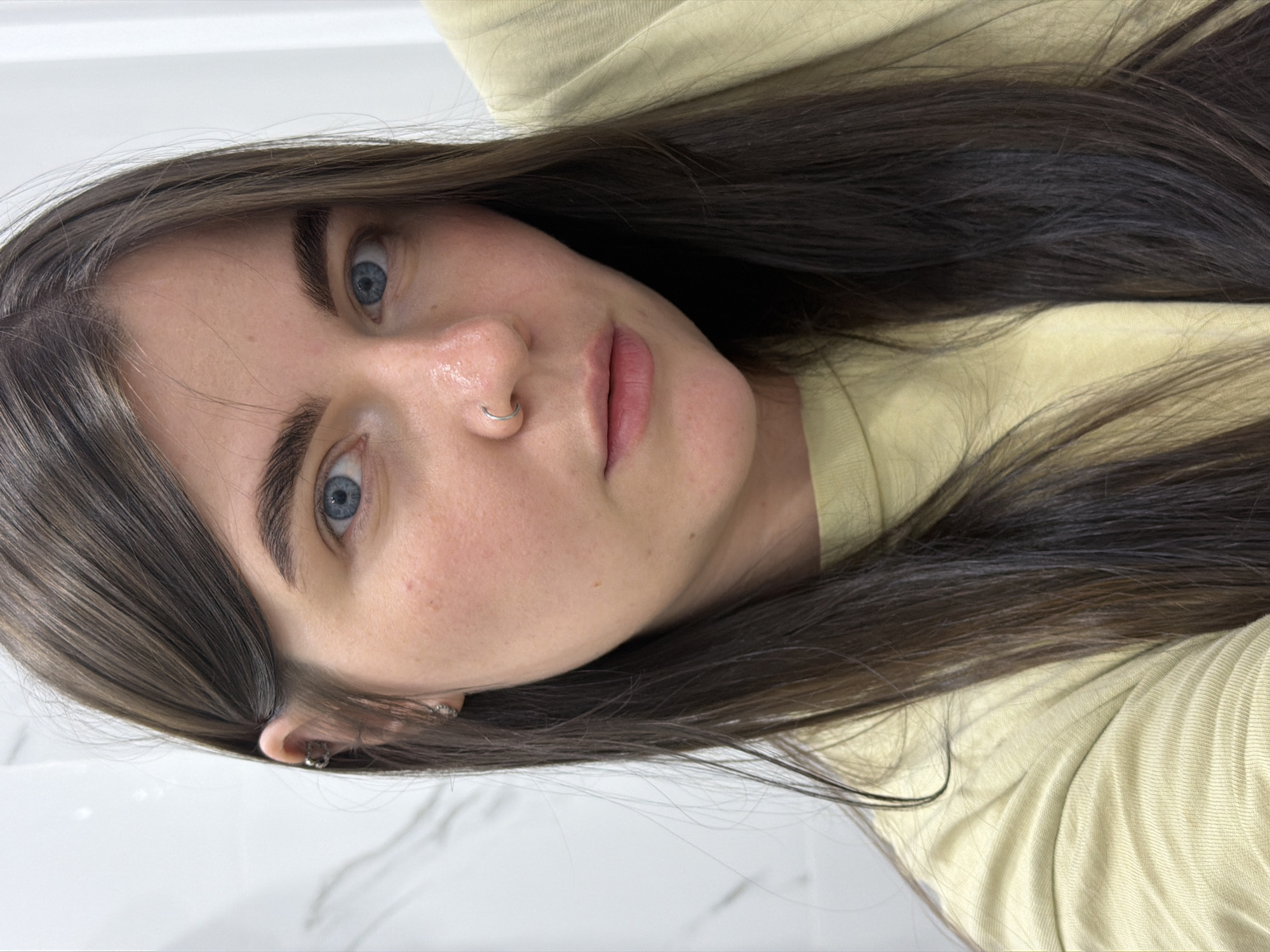 A medium close-up shot presents a woman with light skin and blue eyes, centered in the frame. She has dark hair with highlights and is wearing a light yellow or beige top. The woman is wearing a ring piercing in her nose and a multiple ring earing in her left ear. The background appears to be a flat surface, possibly a white countertop. There are no indications of the location being in Chaikovsky, Russia.