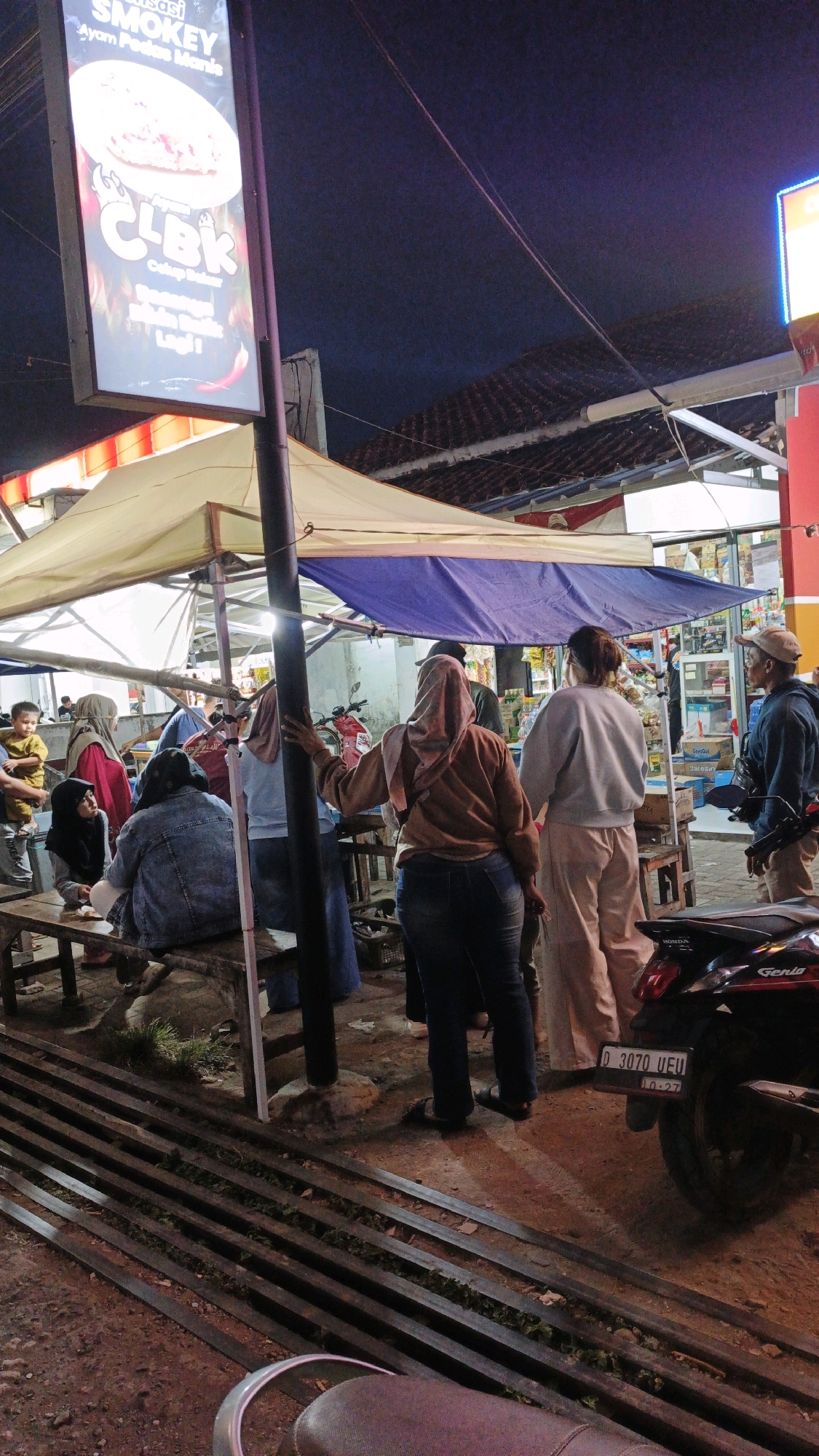 This image depicts a nighttime street scene in Cibenda, Indonesia, likely at a market or food stall area. A prominent sign advertising "SMOKEY AYAM PEDAS MANIS" and "CLBK" is illuminated, suggesting a food vendor. Several people are gathered, some standing and others seated on a wooden bench. They appear to be customers or patrons of the stalls. A tent structure provides shelter overhead, with various goods or merchandise visible in the background, possibly from a small shop. The ground is uneven and appears to be paved with dirt and some metallic grating. A motorcycle is parked on the right side of the frame. The overall atmosphere suggests a casual, communal gathering place at night.