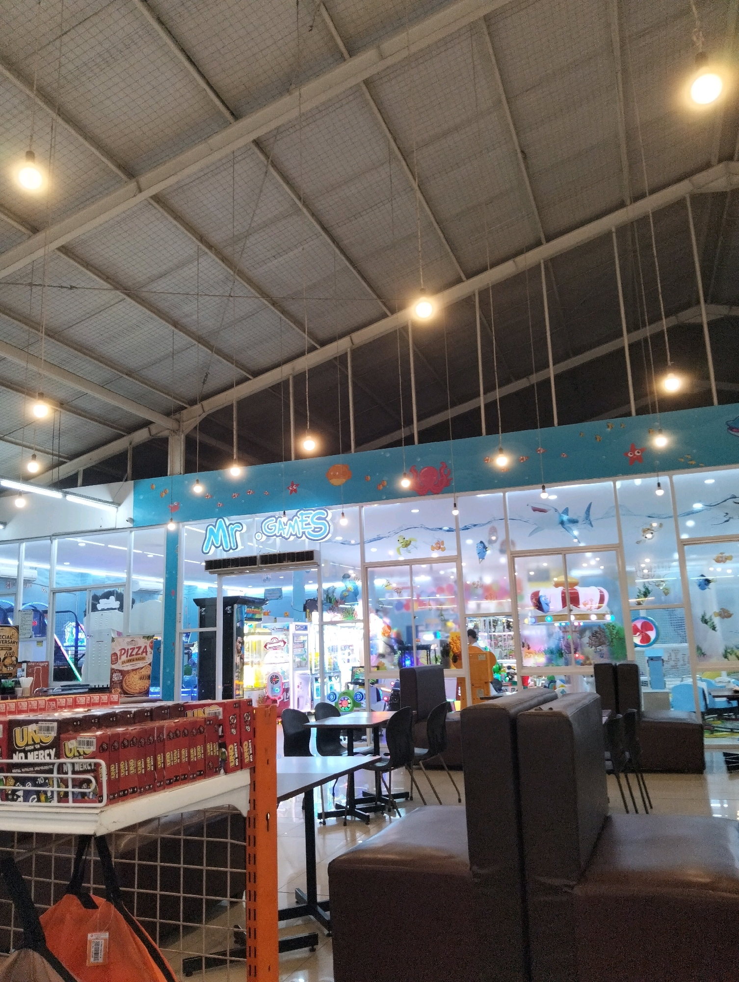 The image captures the interior of an entertainment center, possibly an arcade or indoor playground, identified by the signage "MR. GAMES". The scene is brightly lit by numerous hanging lights and the ambient light from the interior of the establishment. The ceiling structure is visible, featuring a metallic framework with suspended netting. The walls are decorated with a blue underwater theme, complete with cartoonish depictions of marine life like octopuses, fish, and sharks. Large glass windows offer a view into the interior, revealing rows of arcade games, colorful displays, and what appear to be seating areas. In the foreground, there is shelving stocked with boxes of what looks like board games, specifically "UNO NO MERCY". Beyond the shelves, there are tables and chairs, and further inside, more arcade machines and a counter area. The overall impression is one of a lively, family-friendly entertainment venue. The context provided by the user specifies the location as Magelang, Indonesia.
