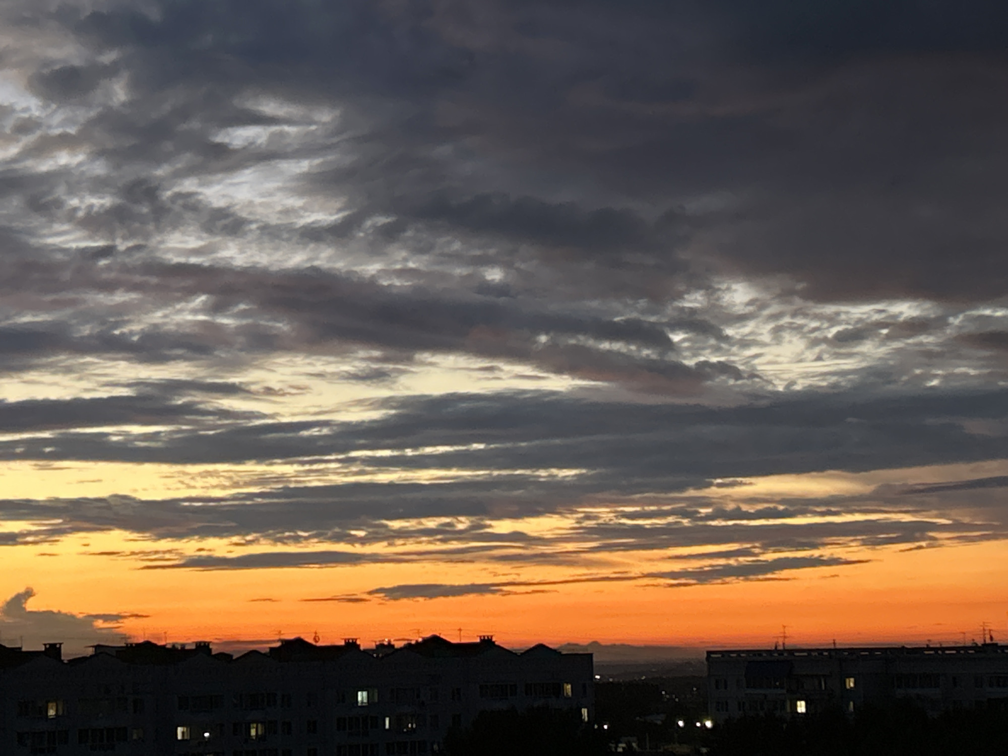The image captures a vibrant sunset over the city of Tomsk, Russia. The sky is a dramatic display of colors, transitioning from a deep, stormy gray at the top to brilliant oranges and yellows closer to the horizon. Wispy, dark clouds are scattered throughout, with some appearing lighter and illuminated by the setting sun. Below the sky, the silhouettes of apartment buildings dominate the foreground, their rooftops and chimneys clearly defined against the colorful sky. Some windows in the buildings are illuminated, suggesting interior lights. The overall scene depicts an urban landscape at dusk, with the natural beauty of the sunset contrasting with the man-made structures.