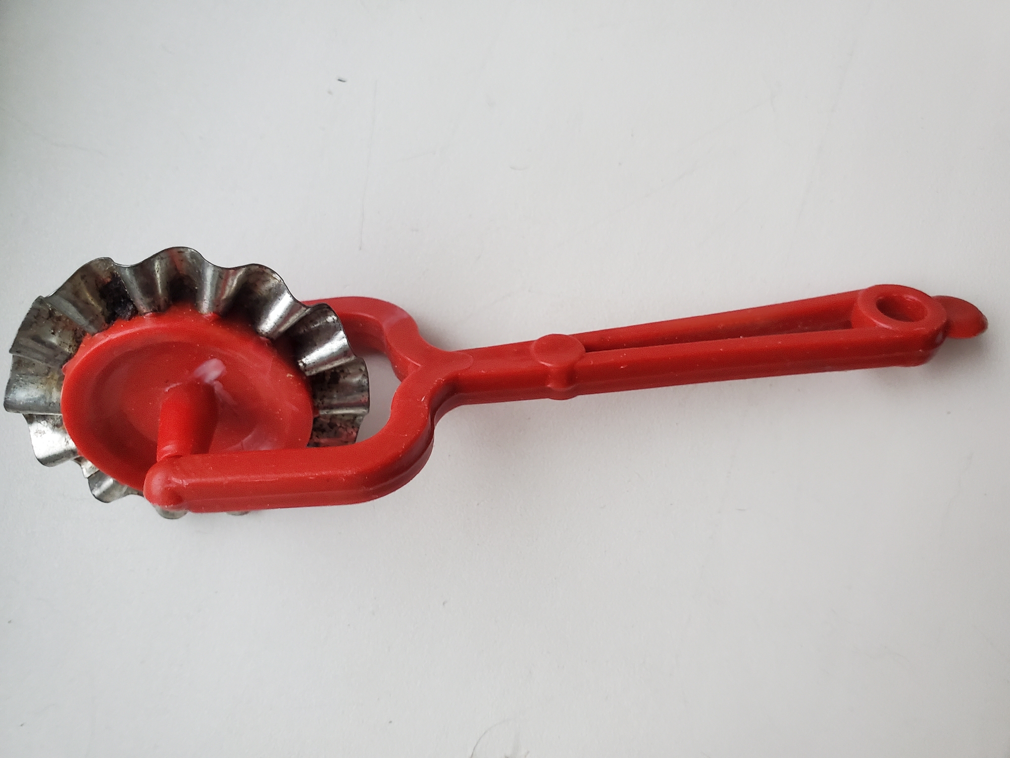 An overhead shot shows a red plastic pastry wheel with a fluted metal cutting edge against a white background. The pastry wheel is positioned vertically in the frame, with the cutting wheel at the top and the handle extending downwards. The red plastic handle has a simple, ergonomic design with a hole at the end, likely for hanging. The metal cutting edge appears slightly tarnished. The background is a plain white surface with subtle textural variations, suggesting it might be a wall or a table. The lighting is even, casting minimal shadows. No people, animals, or other objects are present in the image. The context of Tiraspol, Moldova, is not evident from the visual information provided in the image.