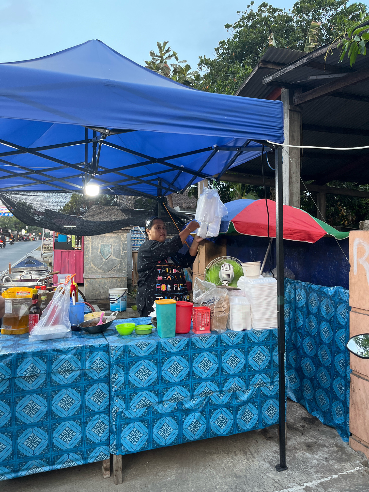 This image depicts a street food stall in Karangpelem, Indonesia. The stall is covered by a large blue canopy, under which a woman is arranging plastic bags. The table in front of her is draped with a blue tablecloth featuring a geometric pattern. Various food service items are visible on the table, including plastic cups, containers, bottles of condiments, and a frying pan. Behind the table, there's a stack of white disposable containers, a green and red umbrella, and a small electric fan. The background shows a road with vehicles and trees, suggesting an outdoor urban setting. The overall scene captures the daily operations of a local food vendor.