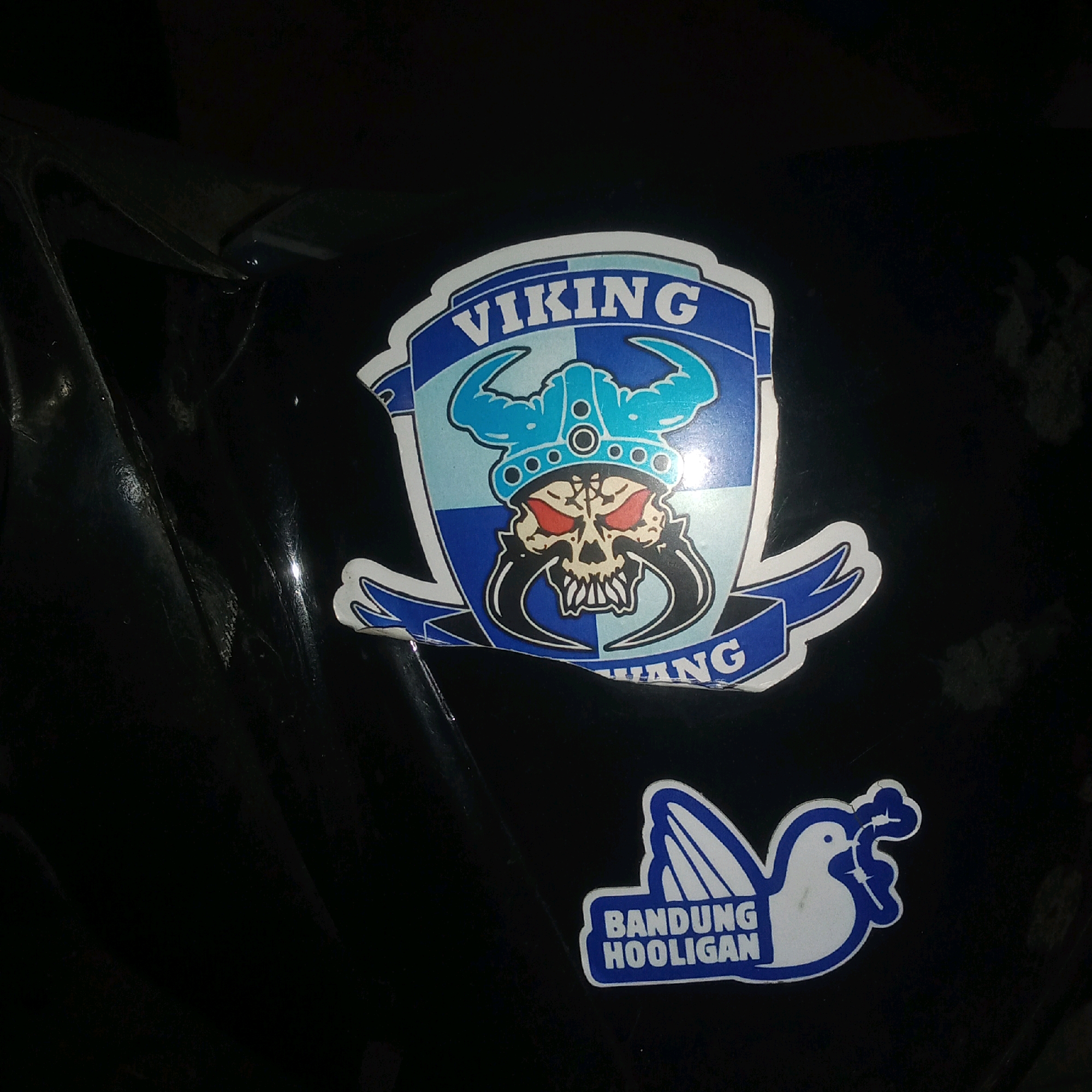 This image displays two stickers on a black, curved surface, possibly a vehicle. The larger sticker, predominantly blue and white, features a Viking helmet on a skull with red eyes and tusks. The word "VIKING" is visible above the helmet, and a partial word "ANG" is seen below it, suggesting "BANDUNG" might be part of the text. The smaller sticker, also blue and white, depicts a stylized bird with outstretched wings and the text "BANDUNG HOOLIGAN" within a blue rectangular border. The context of Karawang, Indonesia, is not directly inferable from the visual elements within the image itself.