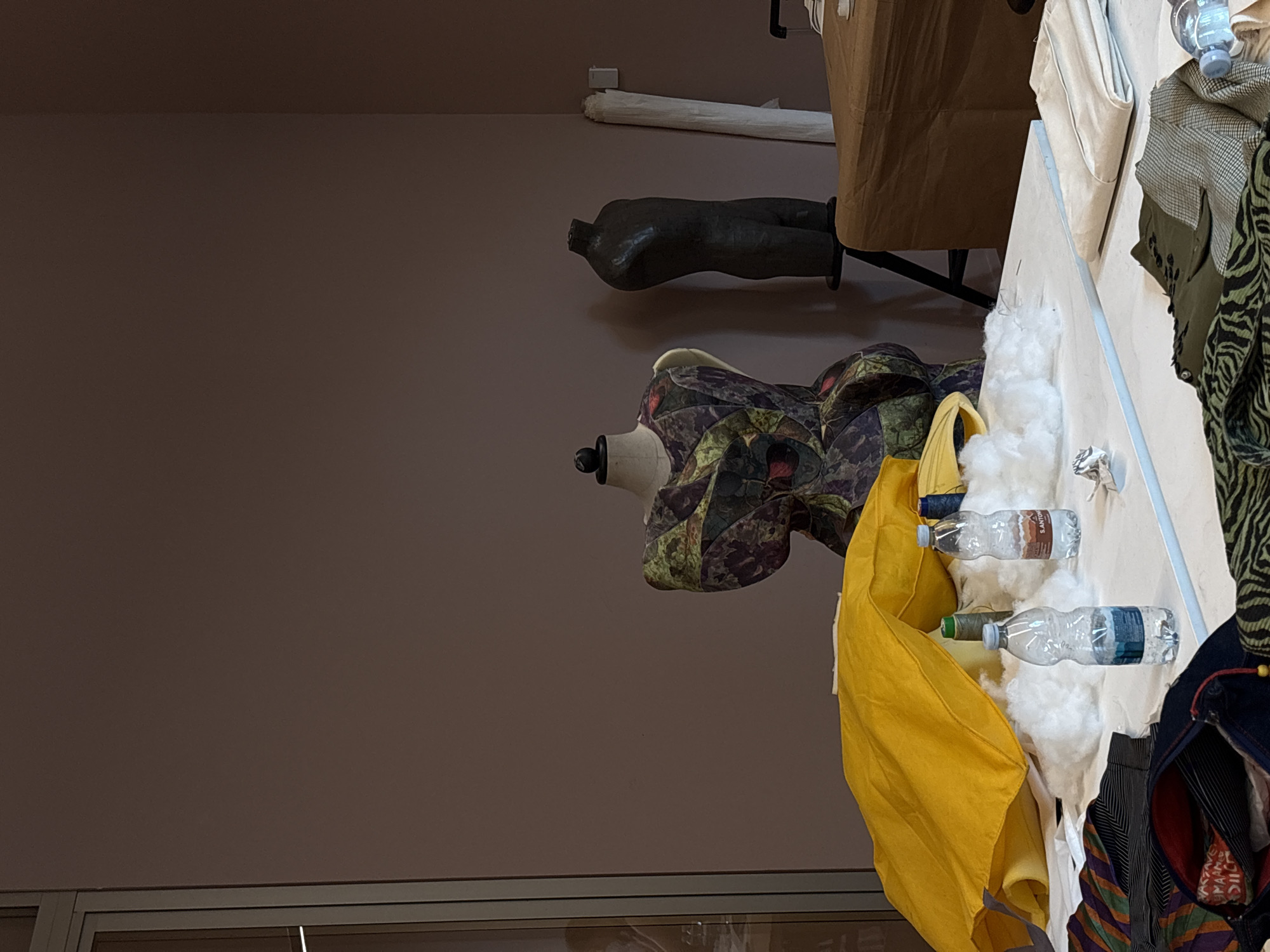 The image shows a mannequin wearing a colorful garment with a pattern of green and purple shapes. The mannequin is positioned on a table covered with fabric scraps, stuffing, and two plastic bottles of water. To the left of the mannequin, there is a dark, stylized torso form, also seemingly made of fabric or plaster. In the background, a roll of white paper or fabric is visible on the floor. The overall setting appears to be a workshop or studio space. The lighting suggests an indoor environment, possibly during the day. The objects suggest a creative process, likely related to fashion design or textile art. No visible text is present in the image.