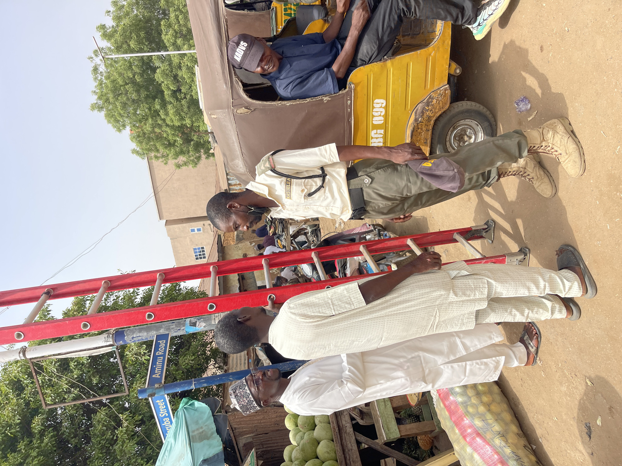 This image captures a bright daylight scene on a street in Zaria, Nigeria, featuring multiple individuals, a vehicle, and market goods.
In the upper-middle, a man wearing a blue shirt, black pants, and a grey cap (with "SAOYS" visible) sits in a weathered yellow auto-rickshaw, often called a Keke Napep, and smiles. The vehicle's side displays "NG 099". Standing next to the rickshaw is a man in a light-colored uniform, consisting of a khaki shirt with "KASTELEA" written on its pocket, olive green trousers, and tan lace-up boots. He wears a dark cap and has a strap across his chest. Above the rickshaw, a person's feet in light-colored sneakers are visible.
To the left, two other men are standing. The one in front wears a white traditional thobe/robe, a white patterned cap, and glasses. Behind him is another man in a cream-colored thobe. A prominent red ladder extends horizontally across the frame, partially obscuring the background. Below the ladder, a blue street sign clearly marks "Aminu Road" and partially "Gariba Street".
The foreground features market items, including fresh green mangoes piled up and a white mesh sack with "HARLEYS" printed on it, likely containing other fruits. The background shows modest buildings, one with distinct blue-framed windows, and green-leafed trees under a clear, bright sky. The strong sunlight and visible shadows suggest it is midday or early afternoon. The scene portrays typical street activity and commerce in a bustling urban environment.