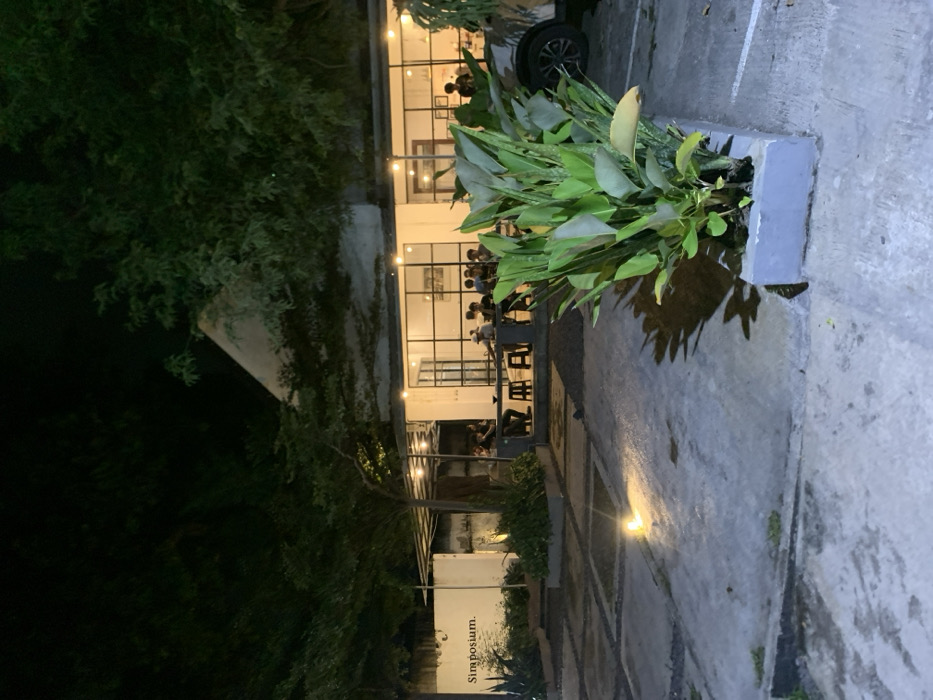 The image shows a night scene of a restaurant with a white exterior and large glass windows. The restaurant is located in Probolinggo, Indonesia, at the coordinates -7.751 latitude and 113.213 longitude. The restaurant is illuminated by warm light, and there are several people seated at tables outside the restaurant, facing inward. The tables are surrounded by a small patio area with black metal chairs. In the foreground, a large potted plant, a dark grey concrete planter with green foliage, is centered in the image. The ground in front of the plant is composed of a light grey, pebbled surface. Behind the plant, there is a dark grey, pebbled surface that extends into the distance. In the background, there is a car parked on the left, only the back left tire is visible. On the right side of the restaurant, there is a white wall with a sign that reads "Simposium." The wall is surrounded by lush green foliage. The scene is dark and peaceful, with the focus on the restaurant and the people inside. The foliage creates a sense of privacy and tranquility.