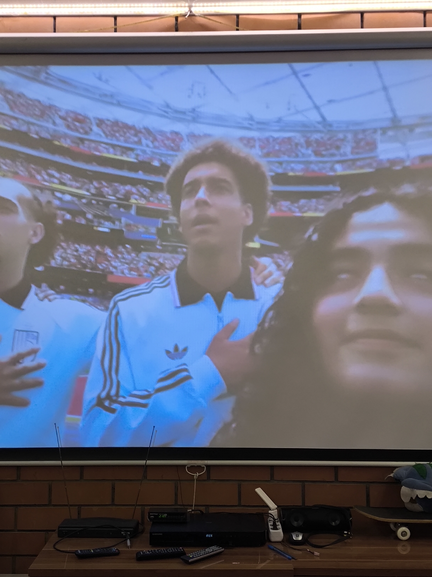 The image depicts a large projection screen displaying a soccer match. In the foreground, on a wooden surface, are several electronic devices, including two remote controls, a black media player with an "AUX" label, and a black soundbar. To the right, a skateboard is visible with a plush toy on it. The background consists of a brick wall. The projection is of a stadium filled with spectators, and three individuals are prominently featured on the screen, two men in athletic wear and one person's face in close-up. One man on the screen is wearing a light blue athletic jacket with dark stripes on the sleeves and an Adidas logo. The location is identified as Pokrov, Russia.