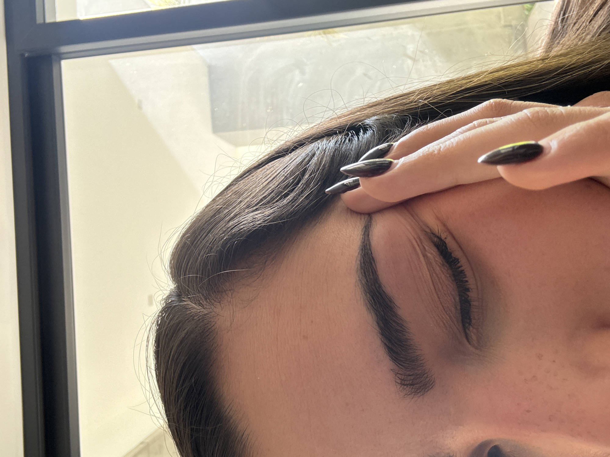 A close-up, oblique-angle shot features a person's face, specifically the forehead, eyebrow, eye, and part of the nose. The person has dark hair and is touching their forehead with fingers that have long, dark-colored fingernails. The background includes a window frame and what appears to be an interior room with light walls. There is no visible information to confirm the location in San Bernardino Tlaxcalancingo, Mexico.
