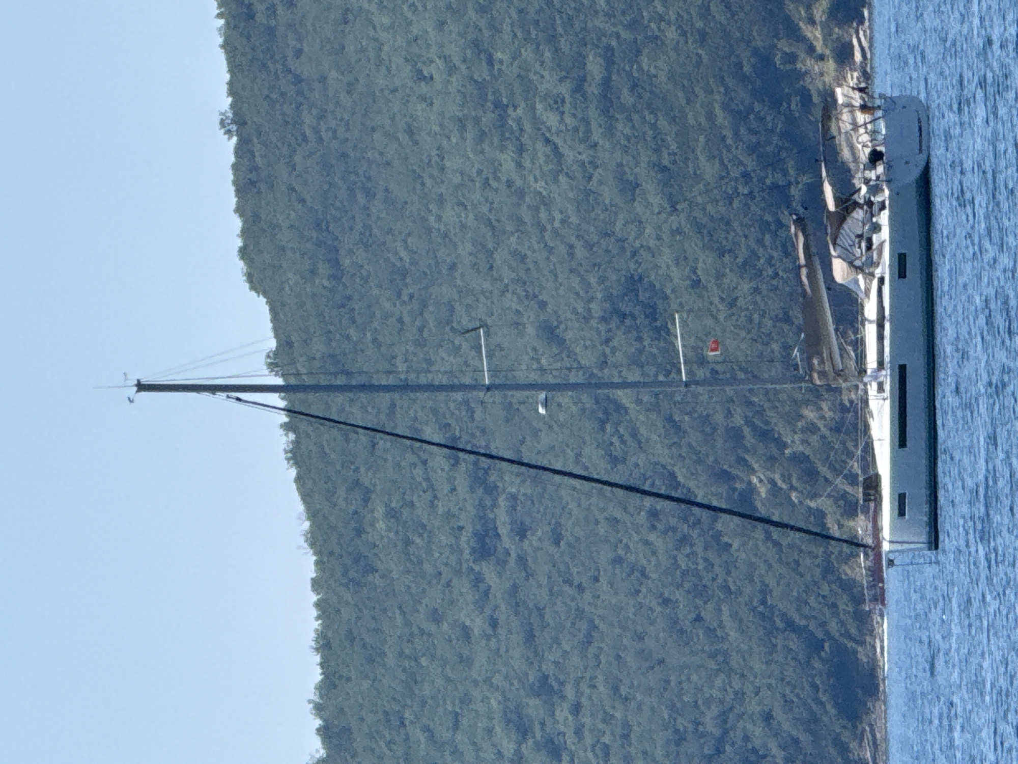 The image shows a sailboat docked in the water near a lush, green hillside. The sailboat is white with dark railings and has a tall mast. The hillside appears to be covered in dense foliage. The water is a deep blue, and the sky is a light blue, suggesting it is daytime and the weather is clear. The scene is tranquil and peaceful, with no people or specific activities clearly visible. The location context provided is Boreti, Montenegro, which is a coastal area known for its natural beauty. There is no visible text in the image.