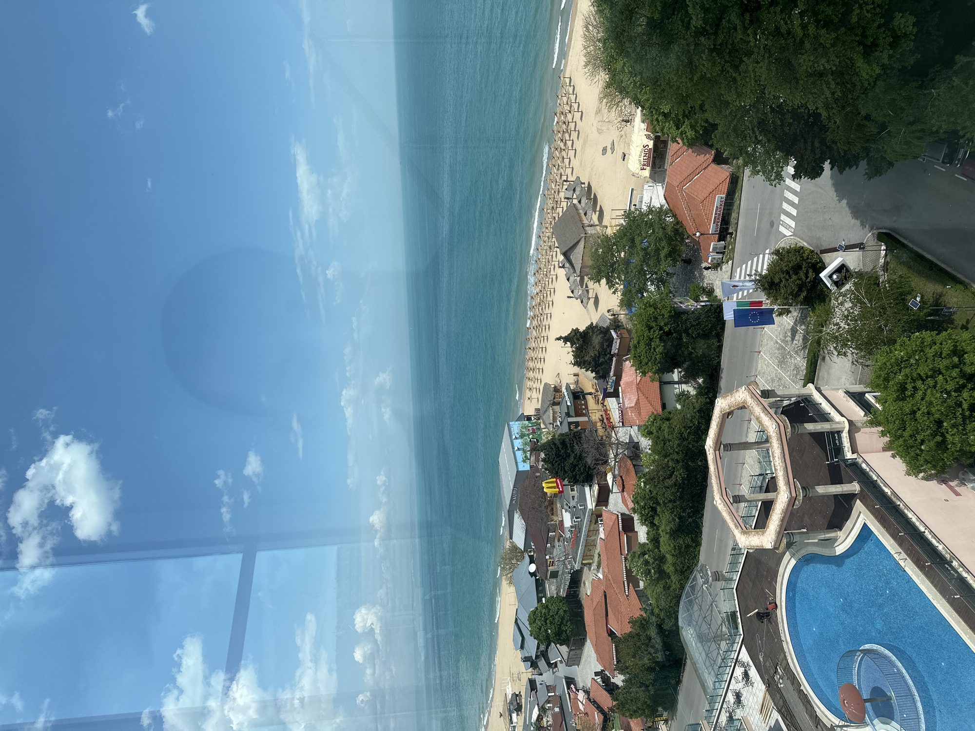 The image presents a high-angle view of a coastal area in Varna, Bulgaria. The scene includes a beach with numerous beach umbrellas and sun loungers, a body of water (likely the Black Sea), and buildings with red-tiled roofs. A road intersects the urban area near the beach. A swimming pool complex is visible, as well as several commercial establishments. The photo appears to be taken from a high vantage point, possibly from a building or observation tower. The sky is partially obscured by what seems to be a window with reflections. National flags of Bulgaria and the European Union are visible in the foreground.
