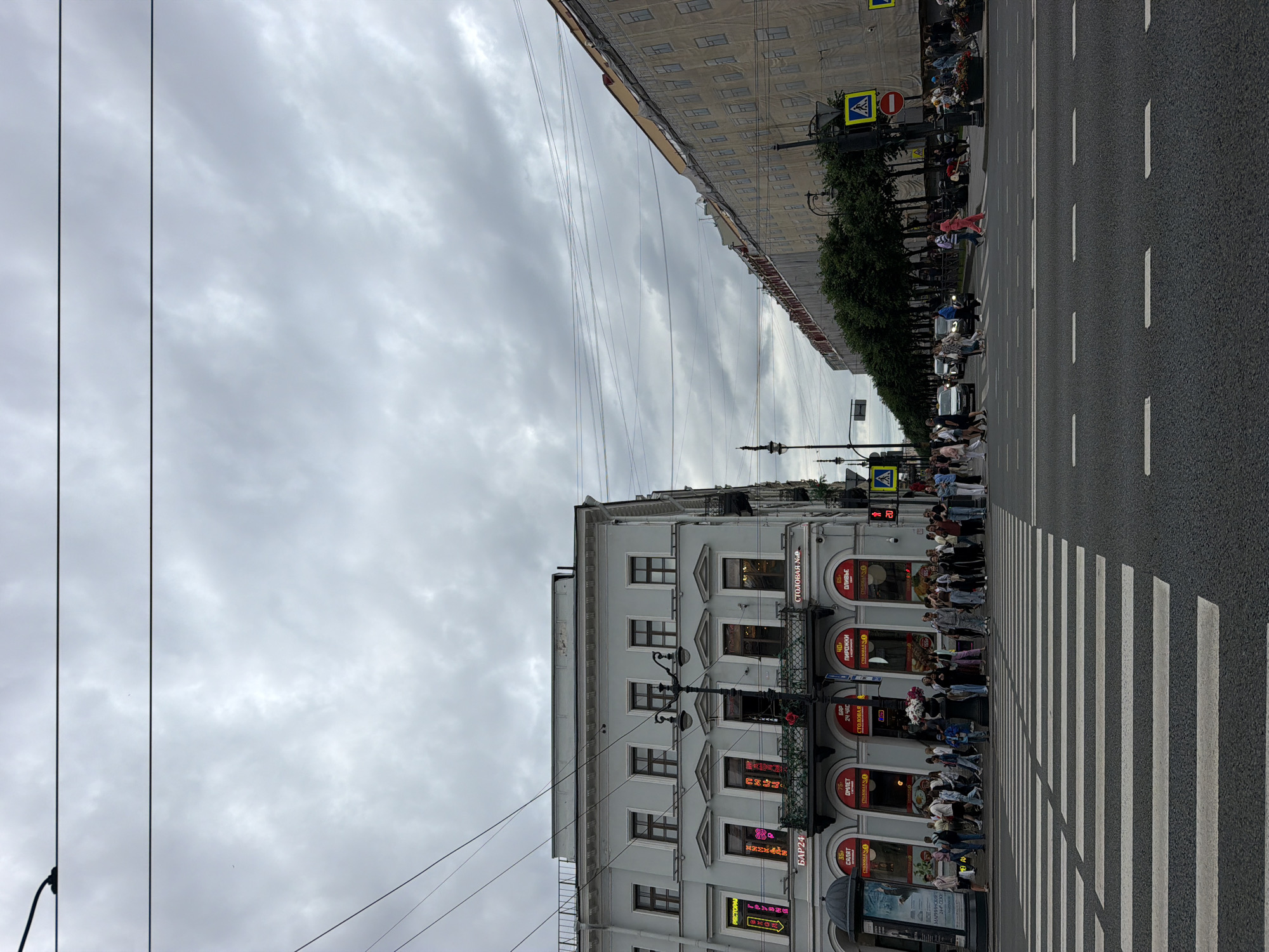 The image captures a street scene in Saint Petersburg, Russia, likely on Nevsky Prospekt given the architectural style and general ambiance. The sky is overcast with heavy grey clouds. In the foreground, a wide pedestrian crossing dominates the asphalt road.
On the left side of the street, a light-colored, multi-story building with ornate window frames and balconies lines the street. Several businesses with illuminated signs are visible on the ground floor, including "РЕСТОРАН", "БАР24", and what appears to be "СТОЛОВАЯ". A black street lamp stands in front of this building. A crowd of people is gathered at the pedestrian crossing, waiting to cross or in the process of doing so. Some individuals are holding flowers.
To the right of the main building, a longer, lighter-colored building extends into the distance. This building is covered in scaffolding and large white sheets, suggesting ongoing construction or renovation. A row of lush green trees runs along the sidewalk in front of this building, creating a visual separation. Street lamps and pedestrian crossing signs are visible along this side of the street. Several cars and people are on the road and sidewalk on the right side as well.
The overall impression is of a busy urban environment on a cloudy day, with a mix of historic architecture and ongoing development.